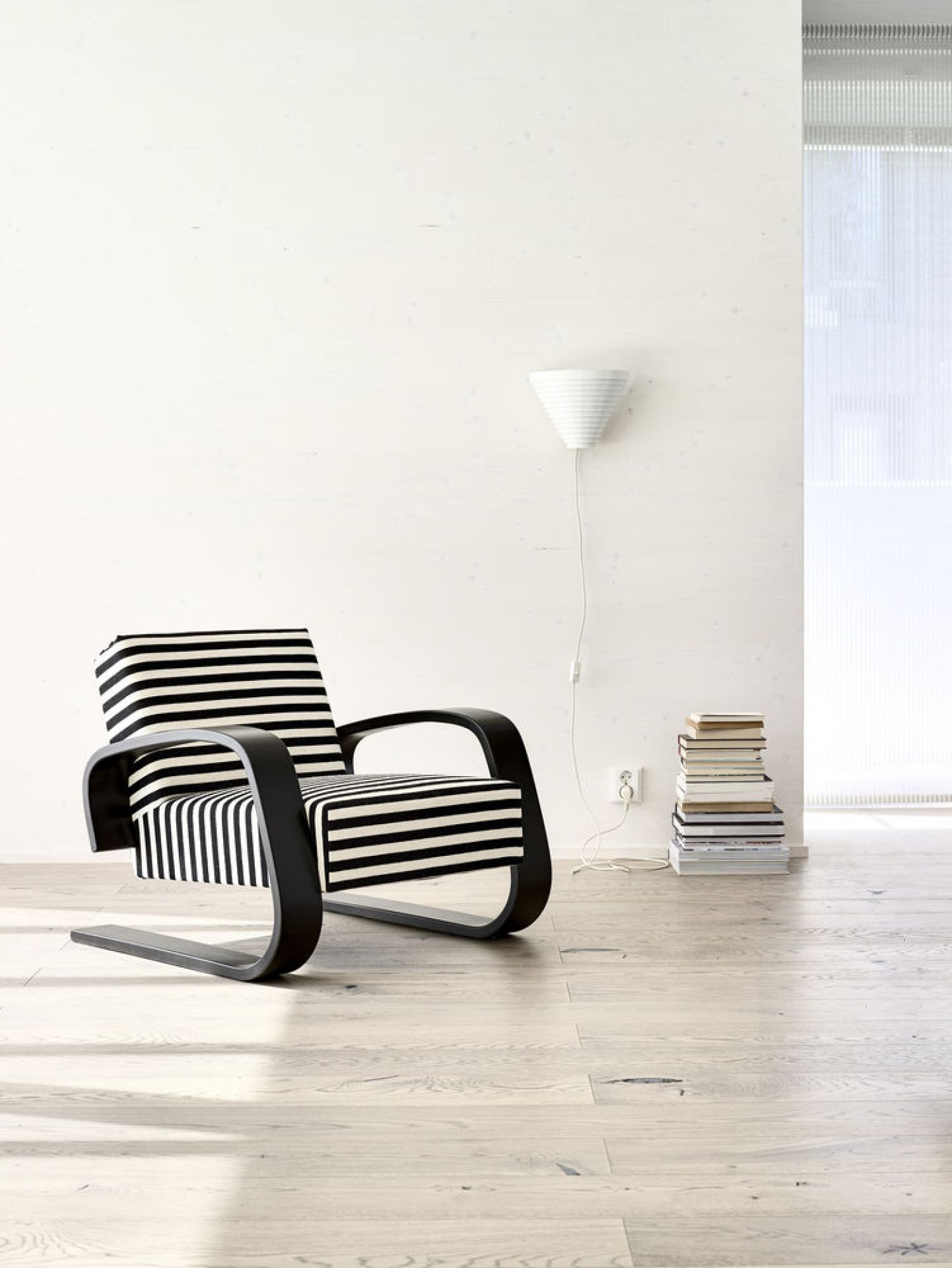 Helle minimalistische Szene mit Artek A910 Wandleuchte, Sessel mit Streifenmuster und Bücherstapel.