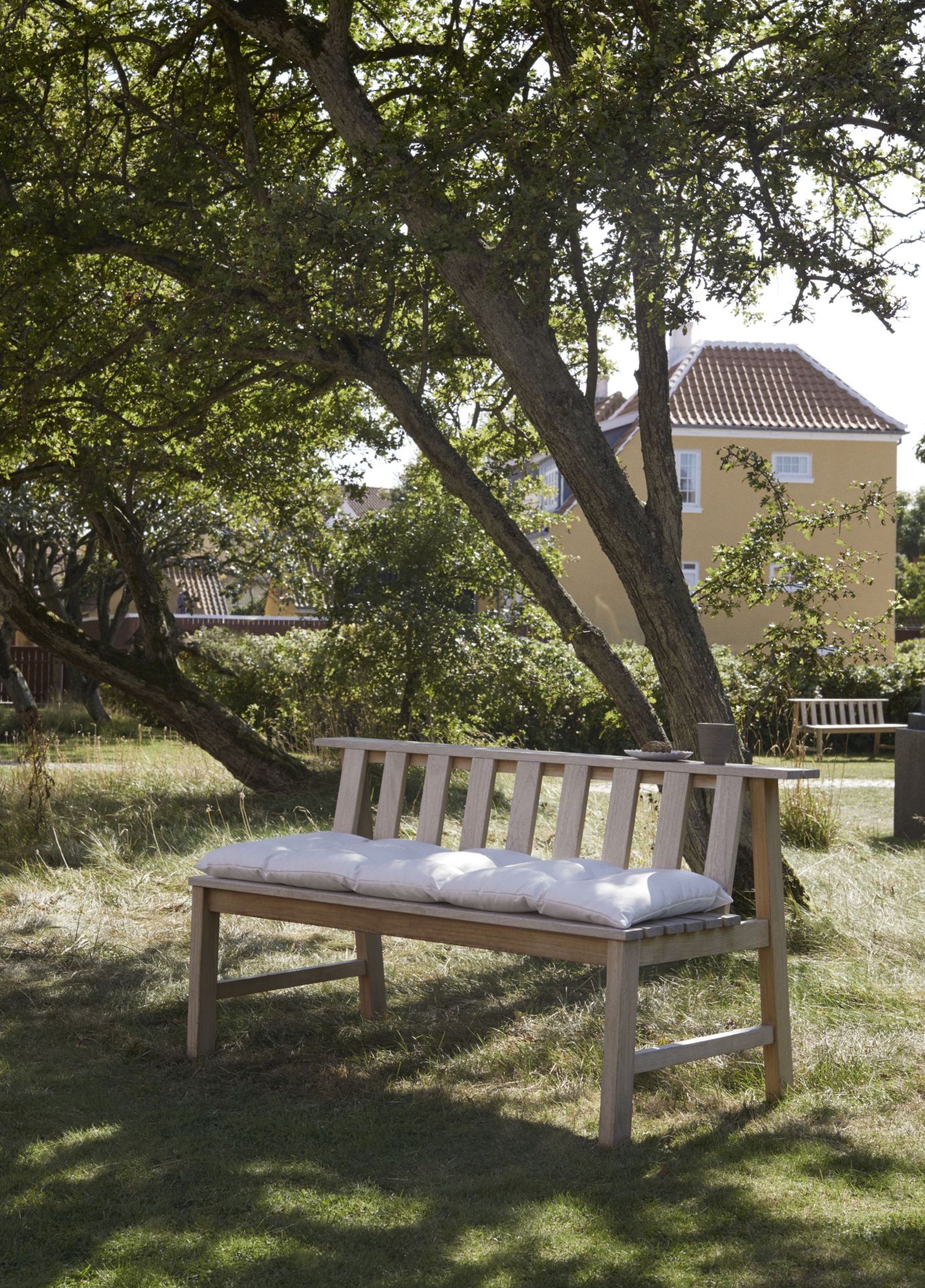 Cadence Teak Gartenbank von Skagerak by Fritz Hansen mit weißen Kissen im sonnigen Garten.