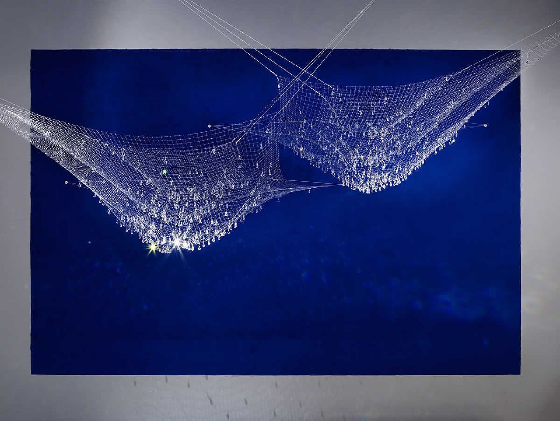 LACRIME DEL PESCATORE Pendelleuchte von Ingo Maurer mit zwei Netzen und tropfenförmigen Kristallen vor blauem Hintergrund.
