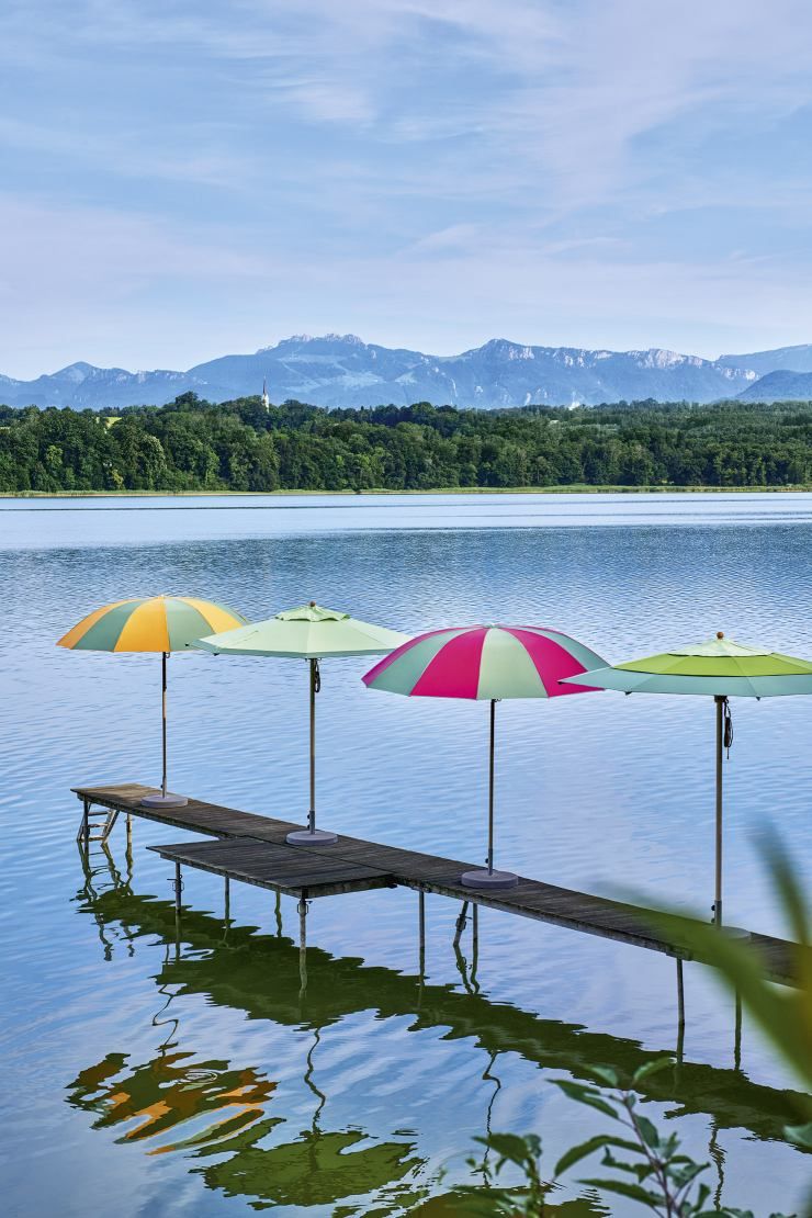 Mehrere bunte Weishäupl Sonnenschirme auf einem Steg im See mit Bergpanorama im Hintergrund.