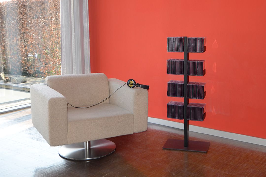 Schwarzer CD-Baum/DVD-Baum Standregal mit CDs neben beigem Sessel und Kopfhörern vor oranger Wand.