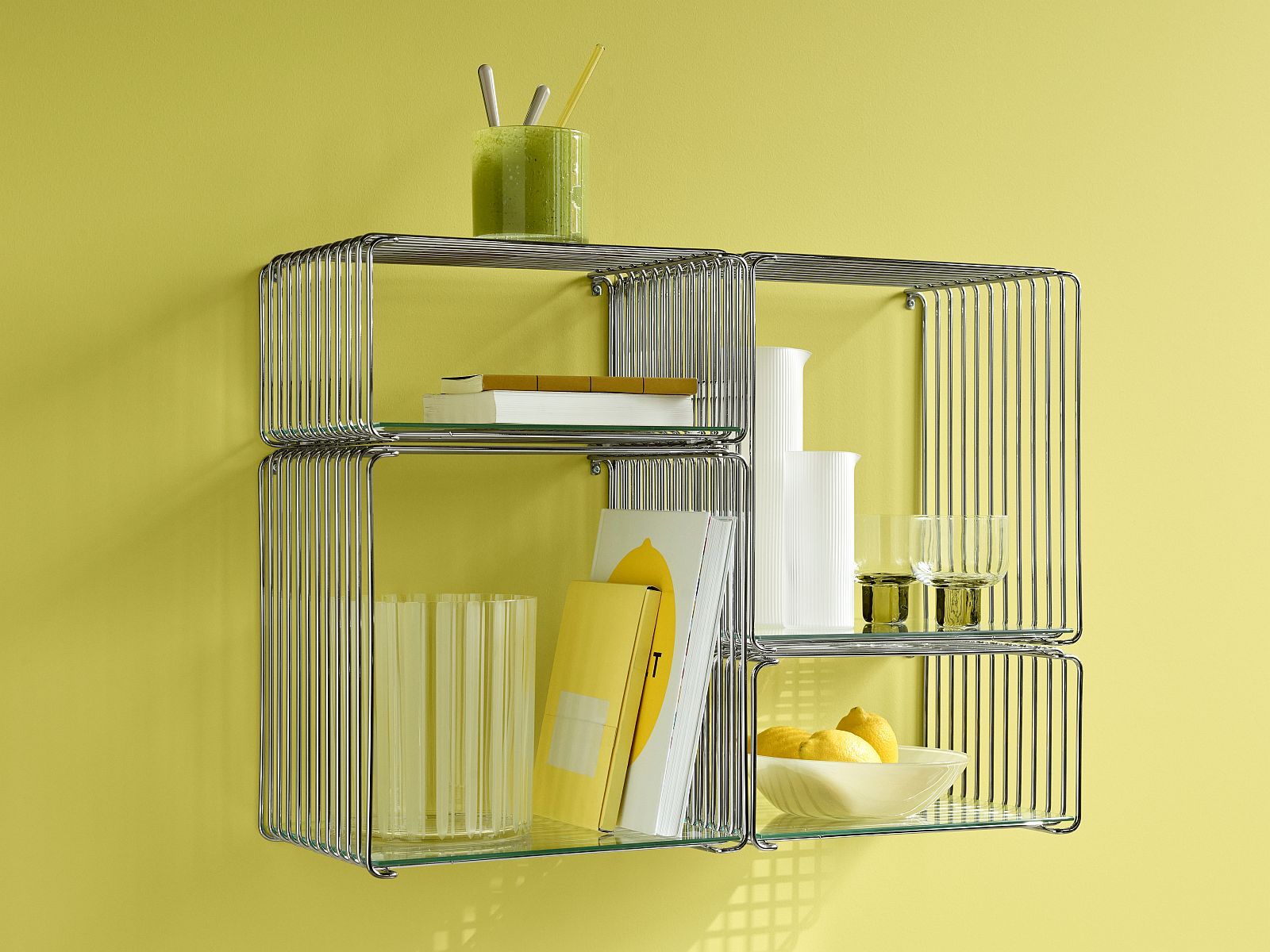 Panton Wire Cube von Montana, verchromtes Drahtregal mit Glasböden und dekorativen Gegenständen vor gelber Wand.