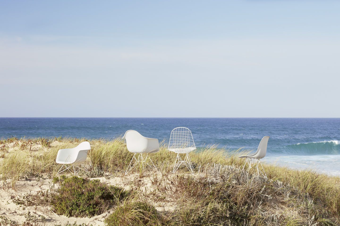 Vier Design-Stühle, darunter ein Vitra Wire Chair, am Strand mit Meer im Hintergrund.