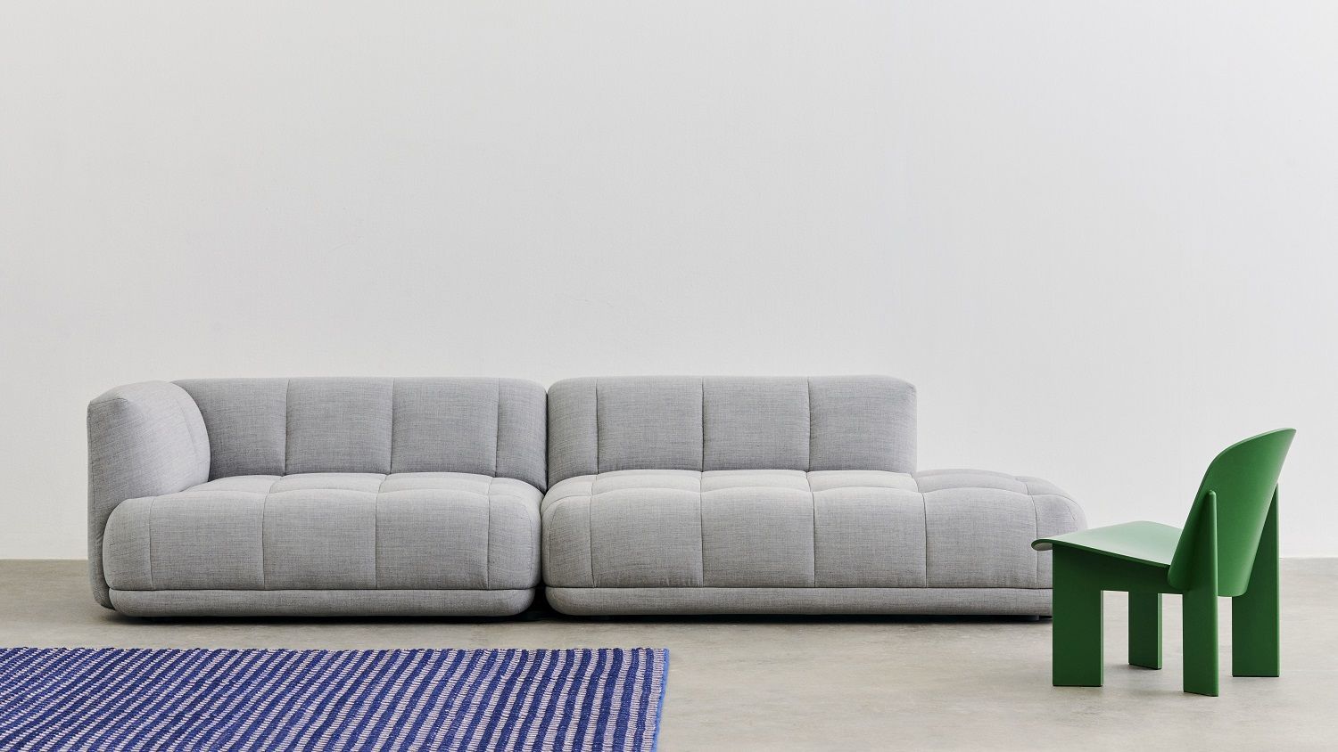 Blau-weißer Channel Rug Teppich von Hay vor grauem Sofa und grünem Stuhl.