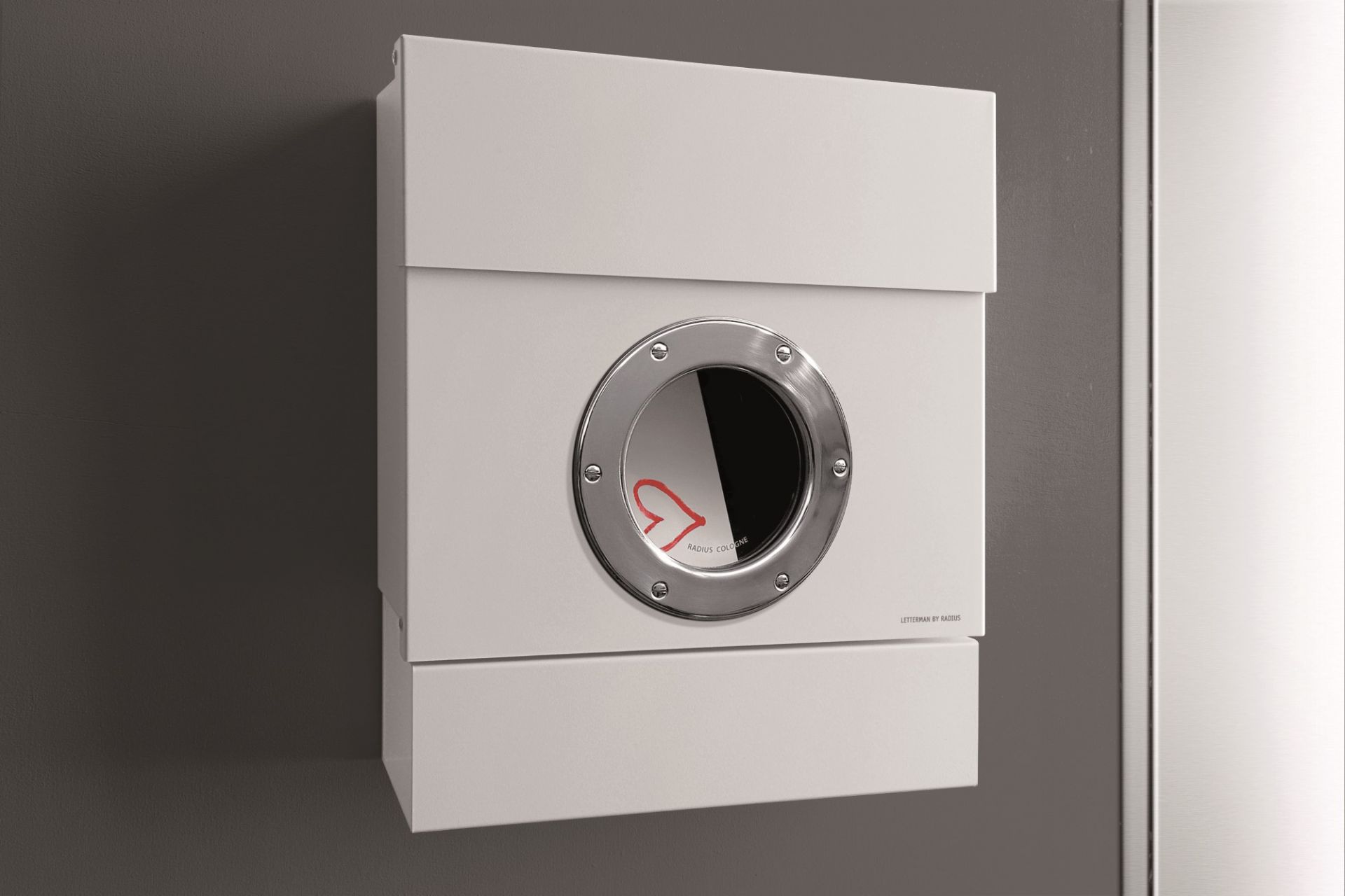 Weißer Letterman Briefkasten Radius II mit rundem Sichtfenster und Edelstahlring an grauer Wand.