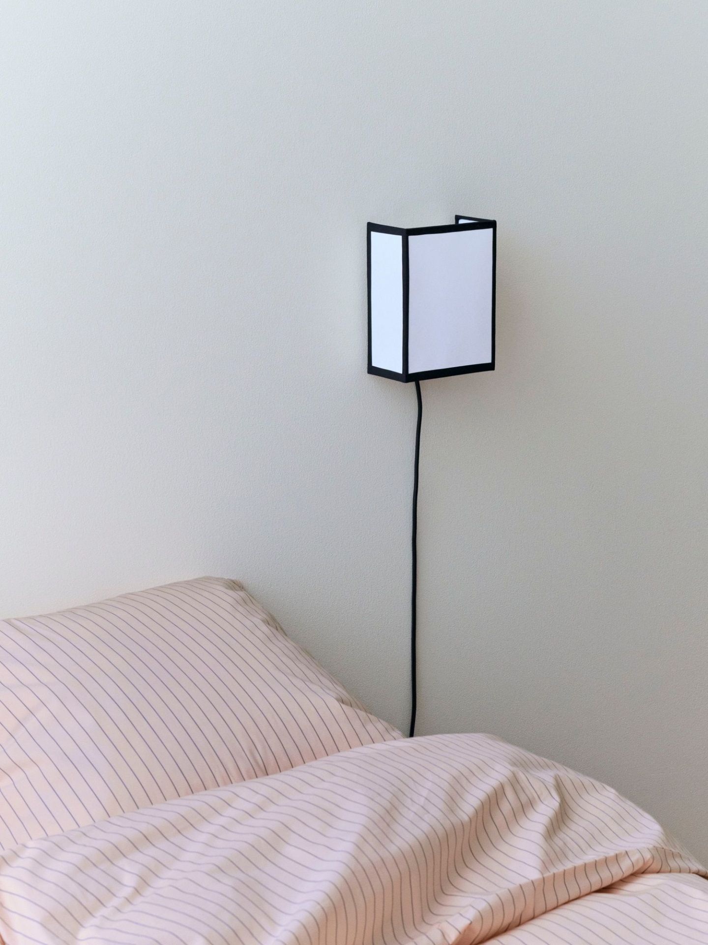 Ava Square Wandleuchte von Hay über einem Bett mit rosa Bettwäsche an einer weißen Wand.