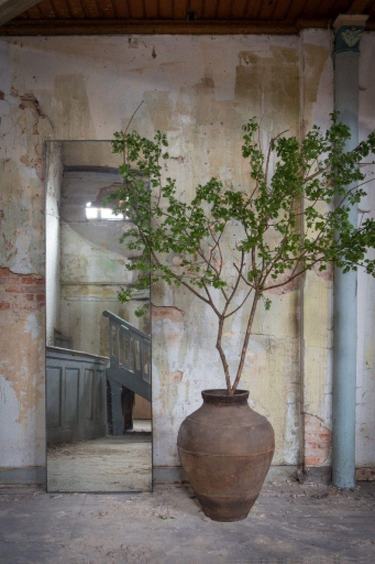 Ethnicraft Wandspiegel "medium aged klar" neben einer Vase mit Zweigen in einem Raum mit Treppe.