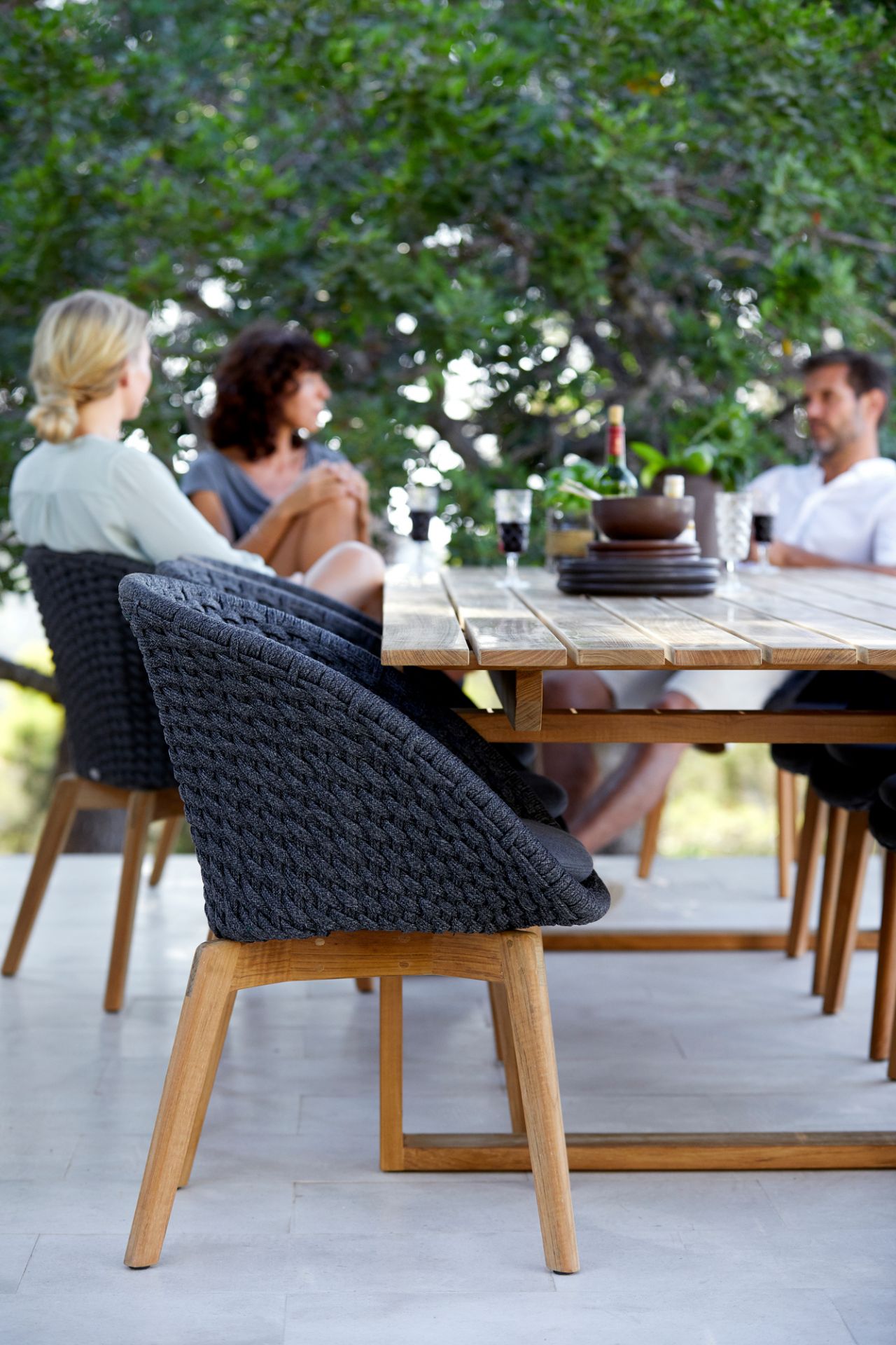 Dunkelgrauer Cane-Line Peacock Outdoor Sessel mit Seilgeflecht und Holzbeinen am Esstisch.
