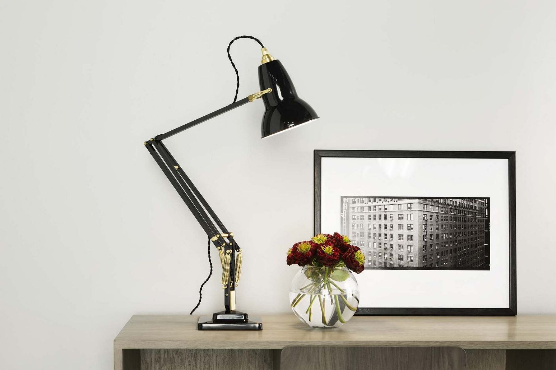 Schwarze Anglepoise Original 1227 Messing Tischleuchte auf einem Schreibtisch, neben einer Vase mit Blumen.
