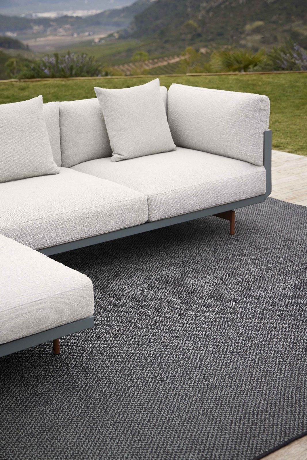 Detailaufnahme des grauen Saline Outdoor Rug Teppichs von Gan unter einem modernen Lounge-Sofa im Außenbereich.