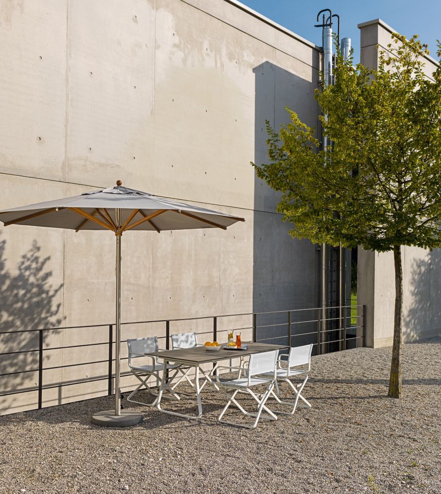 Ahoi Tisch Outdoor von Weishäupl mit Sonnenschirm und vier Stühlen auf Kiesfläche vor Betonwand.