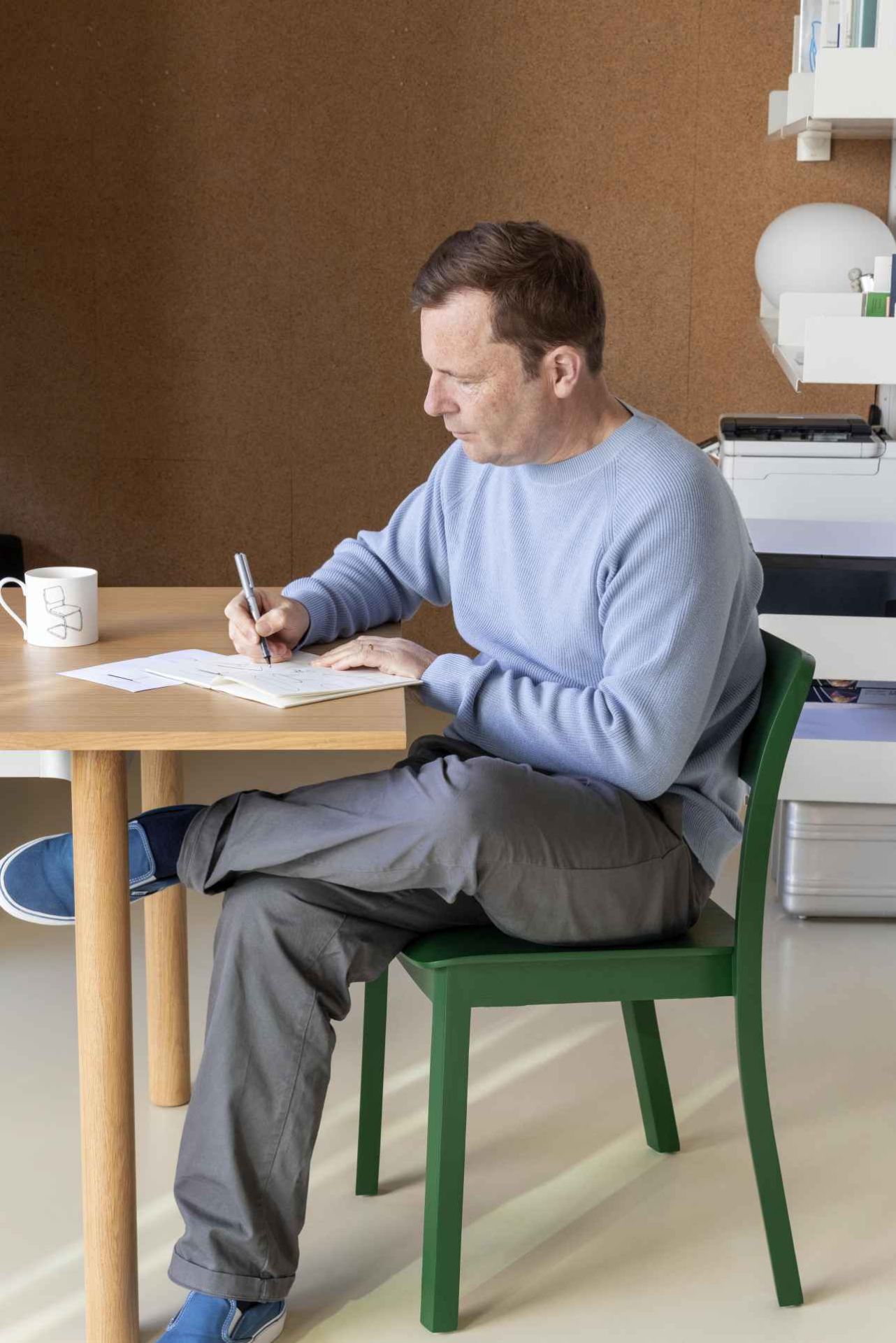 Mann schreibt am Hay Annex Table Esstisch mit hellbrauner Tischplatte und Holzbeinen, dazu ein grüner Stuhl.