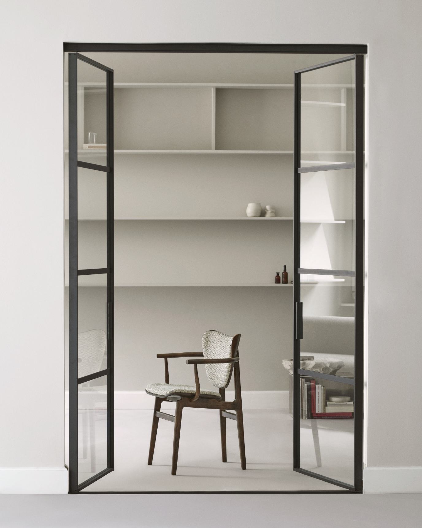 NORR11 Elephant Dining Chair mit Armlehne, Frontpolsterung, Holzbeine, vor Regalen und Glastüren.