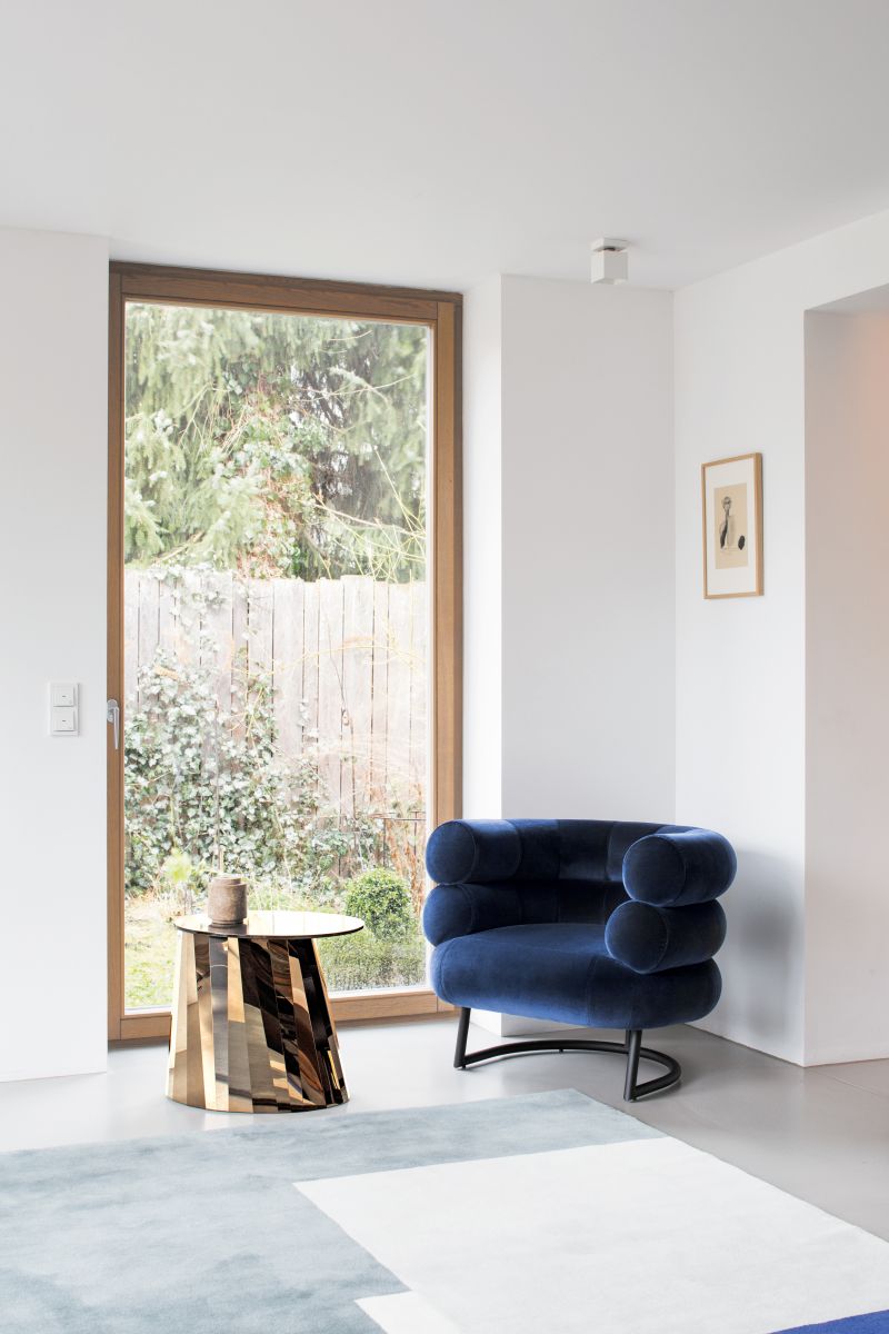 Moderner Wohnraum mit ClassiCon Roquebrune Teppich 300x300cm, blauem Sessel und goldenem Beistelltisch vor Fenster.