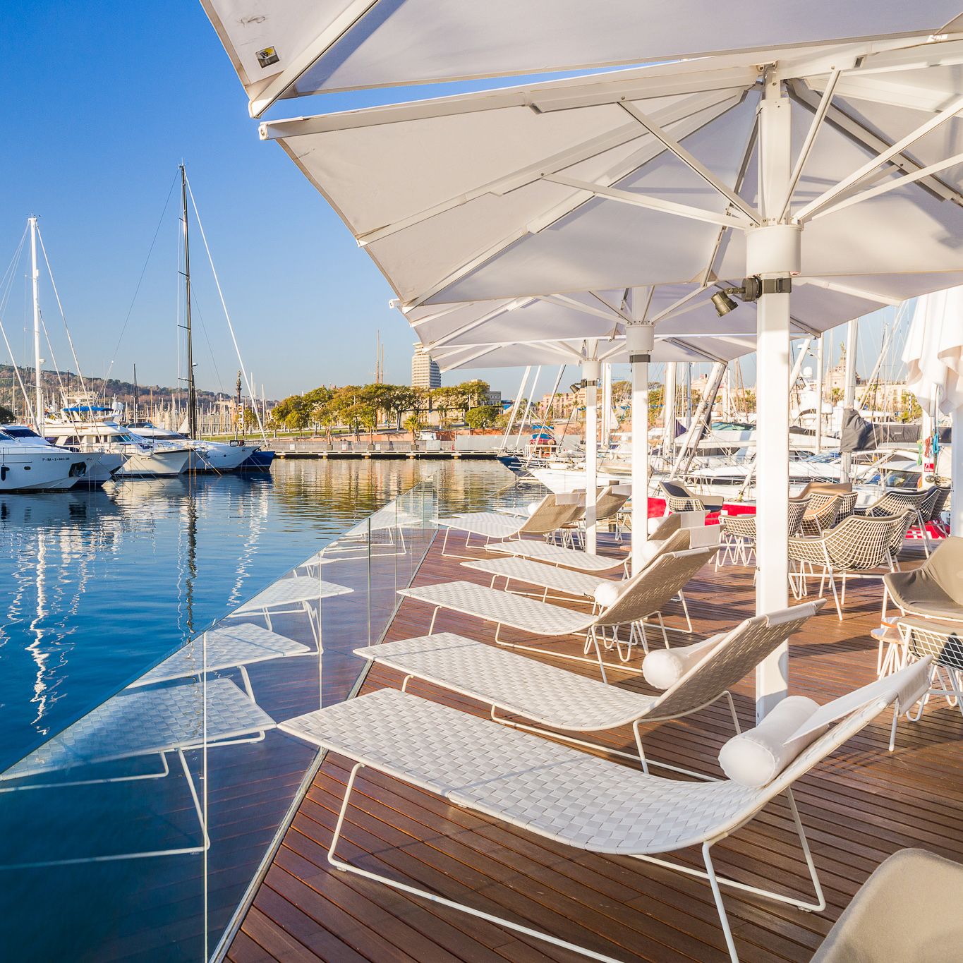 Weiße Trenza Chaise Lounges von Andreu World auf Holzdeck mit Blick auf Yachthafen und Sonnenschirme.
