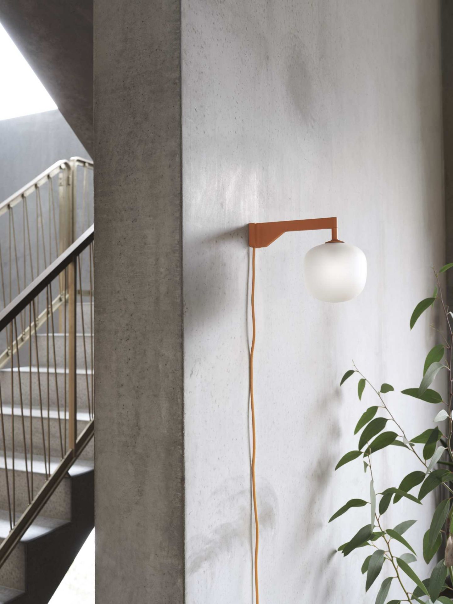 Nahaufnahme der Muuto Rime Wall Wandleuchte mit orangefarbenem Arm und weißem Glasschirm an Betonwand.