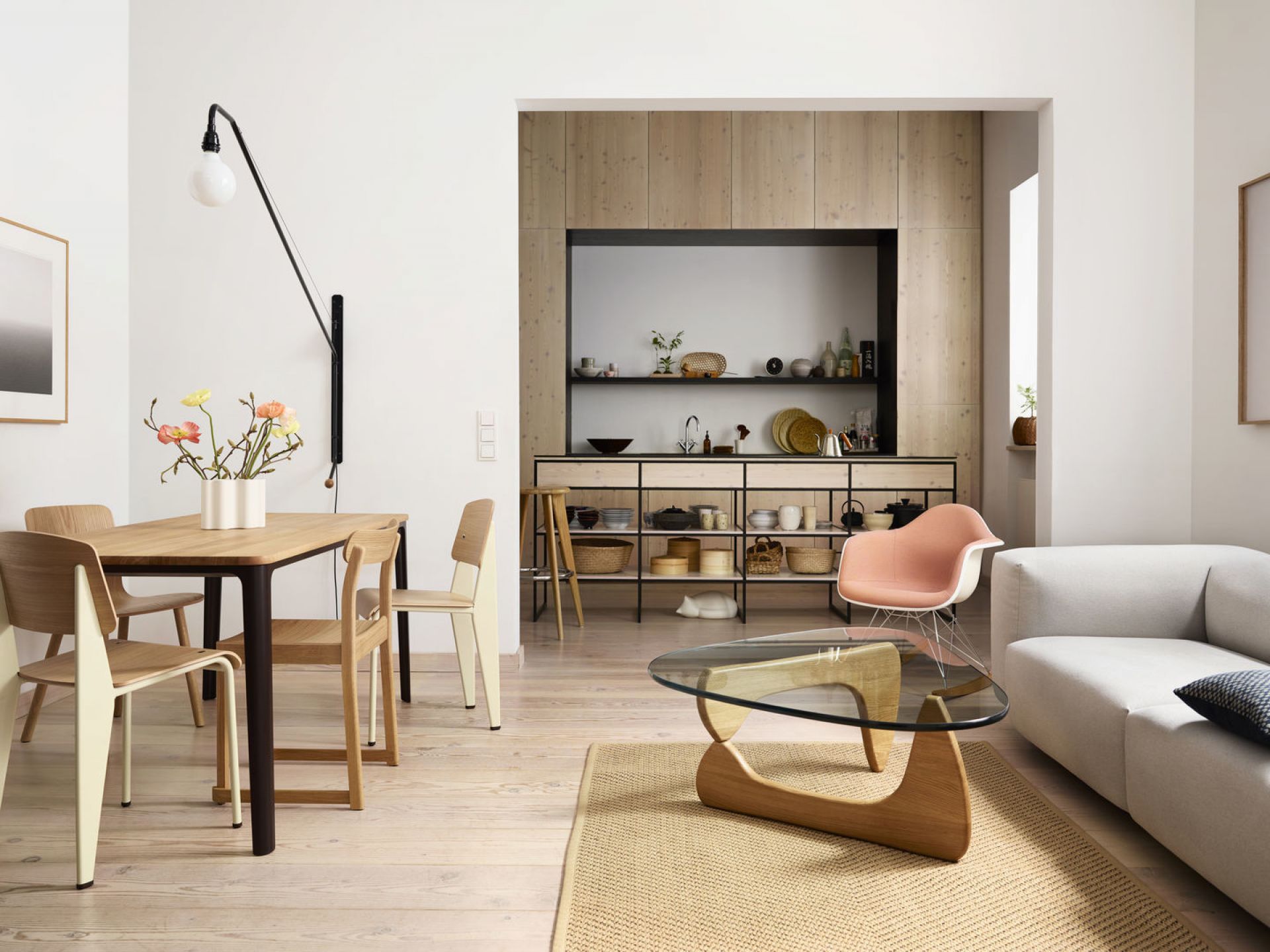 Helles Wohnzimmer mit Esstisch, Holzstühlen, Glas-Couchtisch und offener Küche mit Regalen.