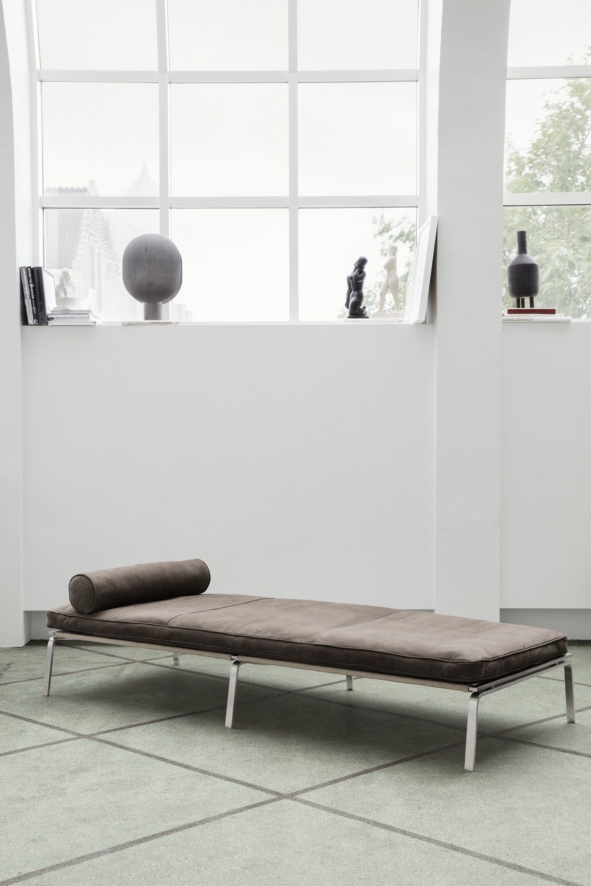 Man Daybed Liege von NORR11 in braunem Leder mit Kissen und silbernen Beinen vor Fenster.