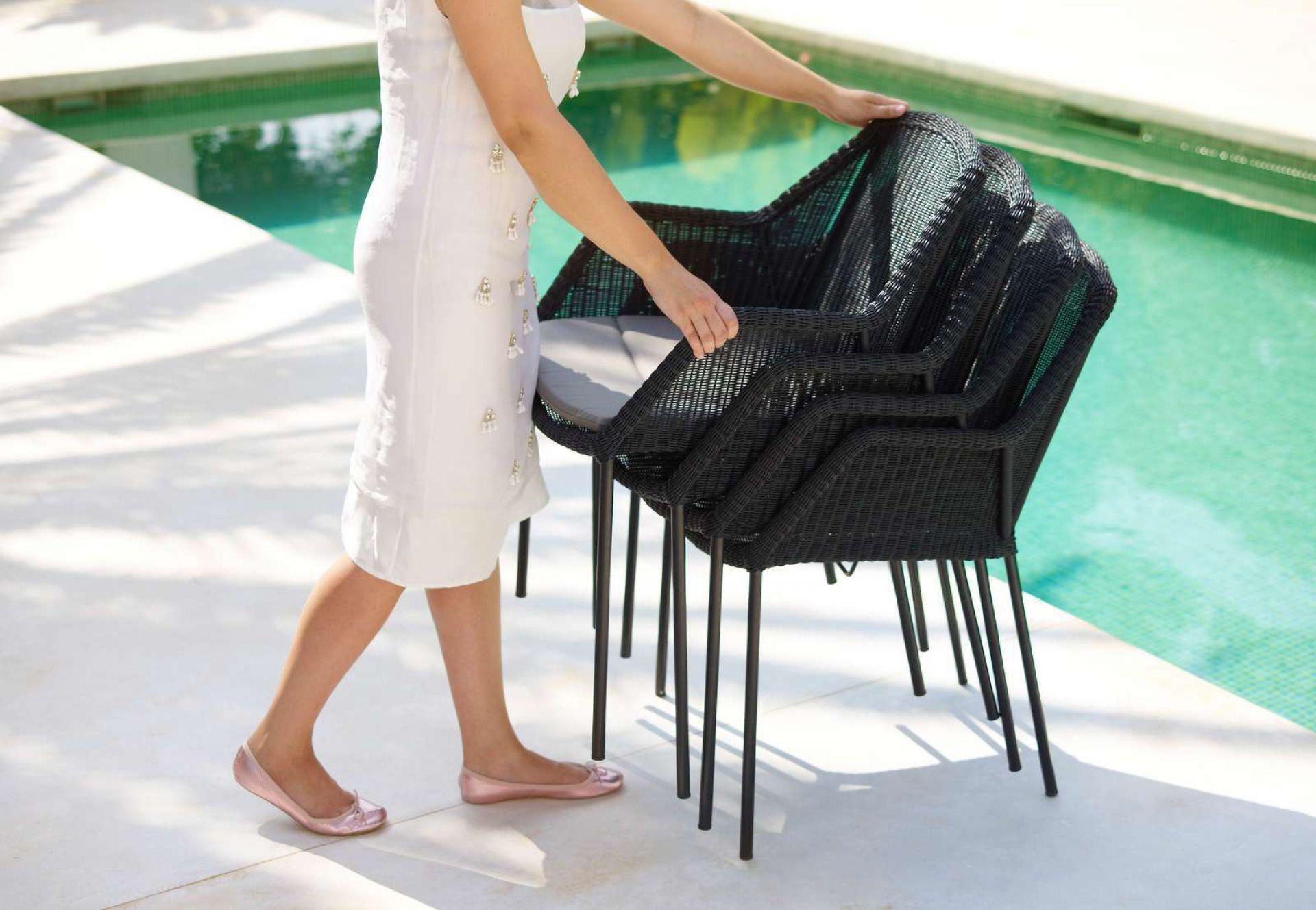Schwarze Cane-Line Breeze Outdoor Stühle mit Vierfußgestell gestapelt, Frau im weißen Kleid daneben, Pool im Hintergrund.