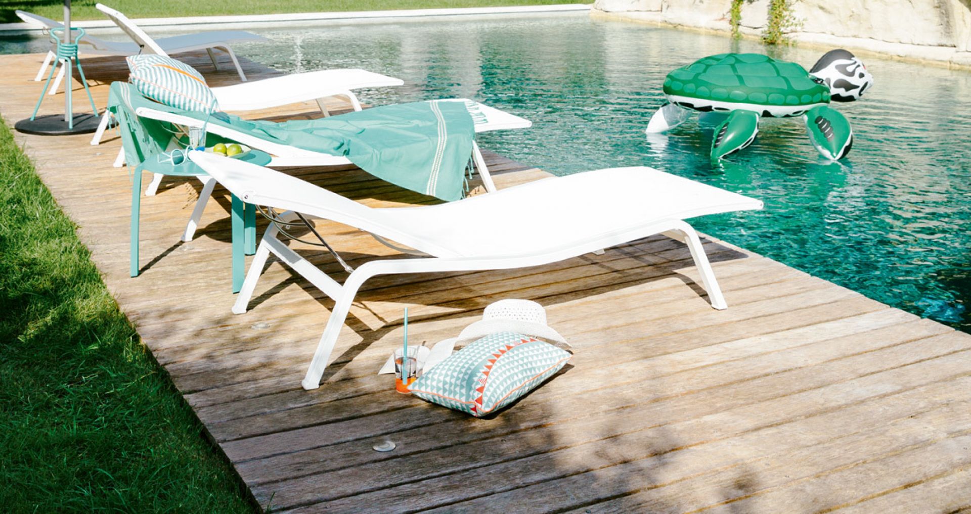 Weiße Alizé Sonnenliege von Fermob am Pool auf Holzdeck mit Dekoration und aufblasbarer Schildkröte.