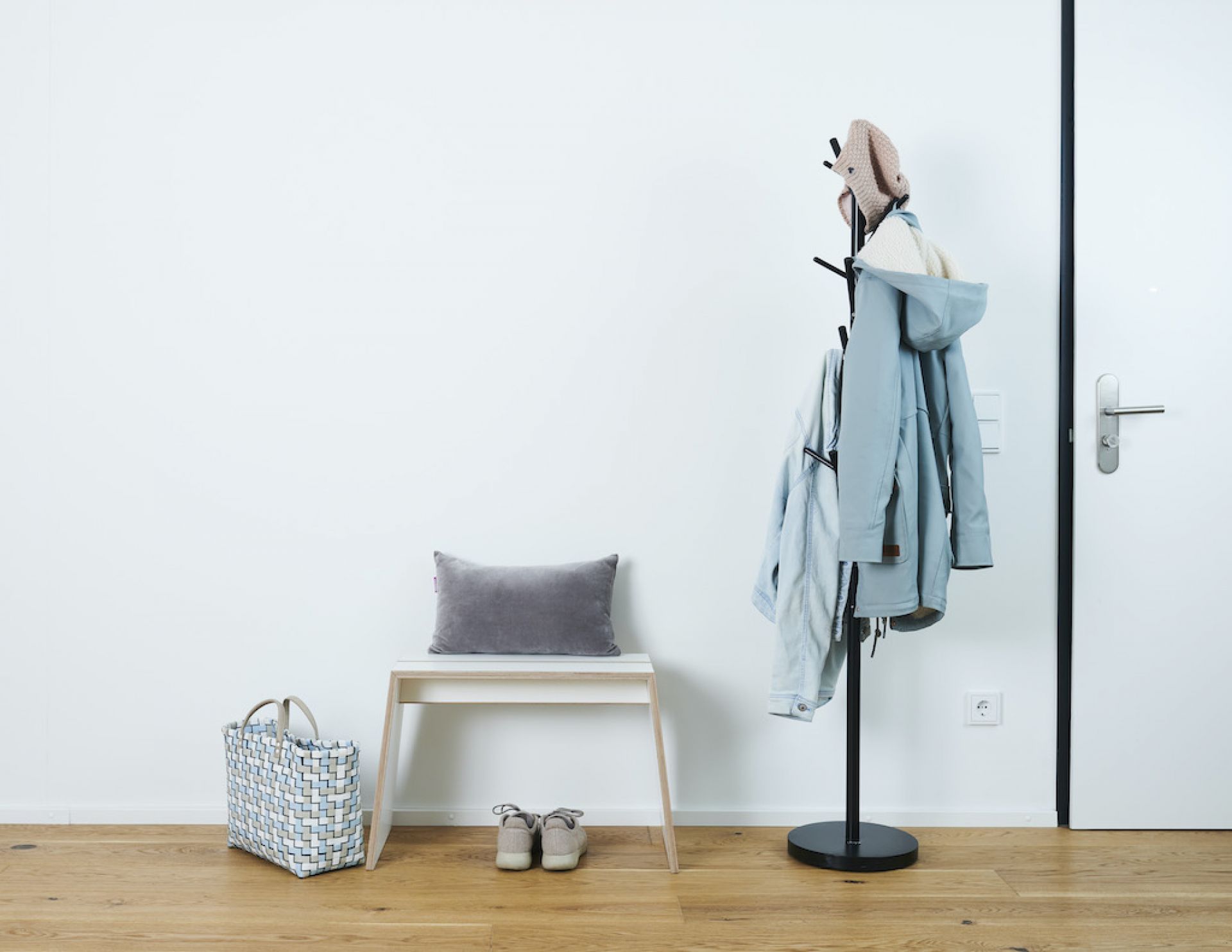 Tojo Stubenhocker Hocker im Flur mit Kissen, Tasche, Schuhen und Kleiderständer vor weißer Wand.