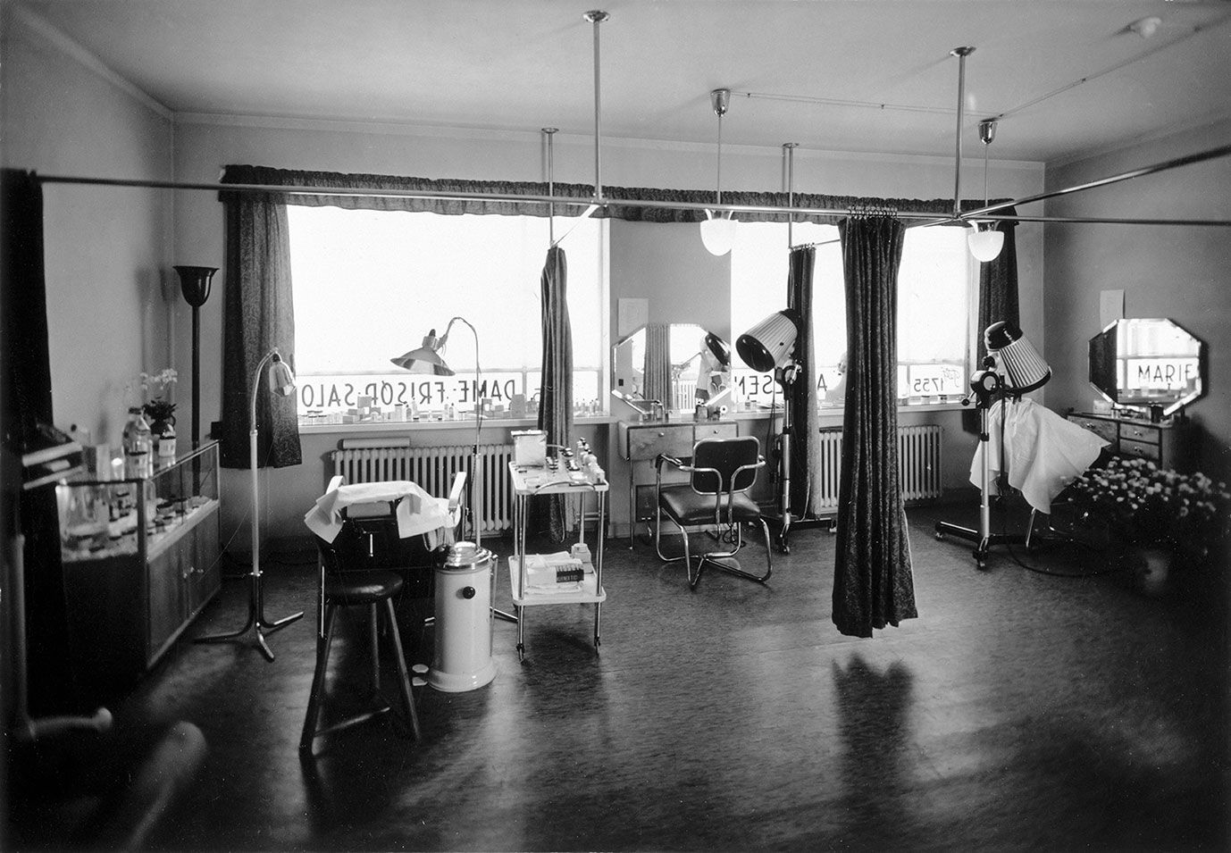 Schwarz-Weiß-Foto eines Friseursalons mit Stühlen, Trockenhauben, Spiegeln und dem Schriftzug "Marie" auf einem Spiegel.