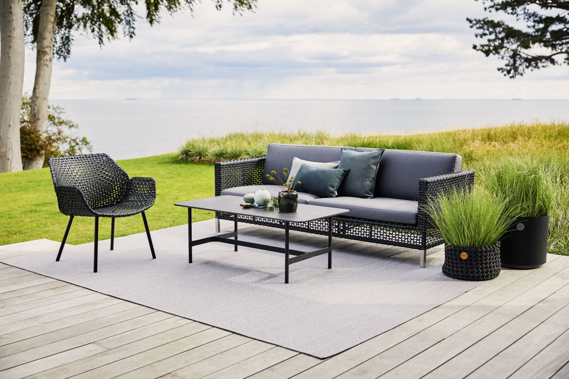 Dot Outdoor Teppich Ø200 cm von Cane-Line auf einer Terrasse mit Gartenmöbeln und Meerblick.