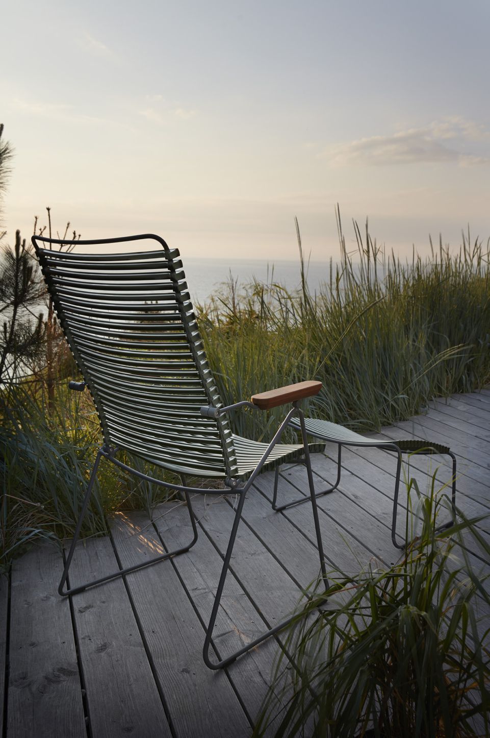 Houe Click Position Chair: Grüner Outdoor Sessel mit Fußhocker auf Holzterrasse am Meer.