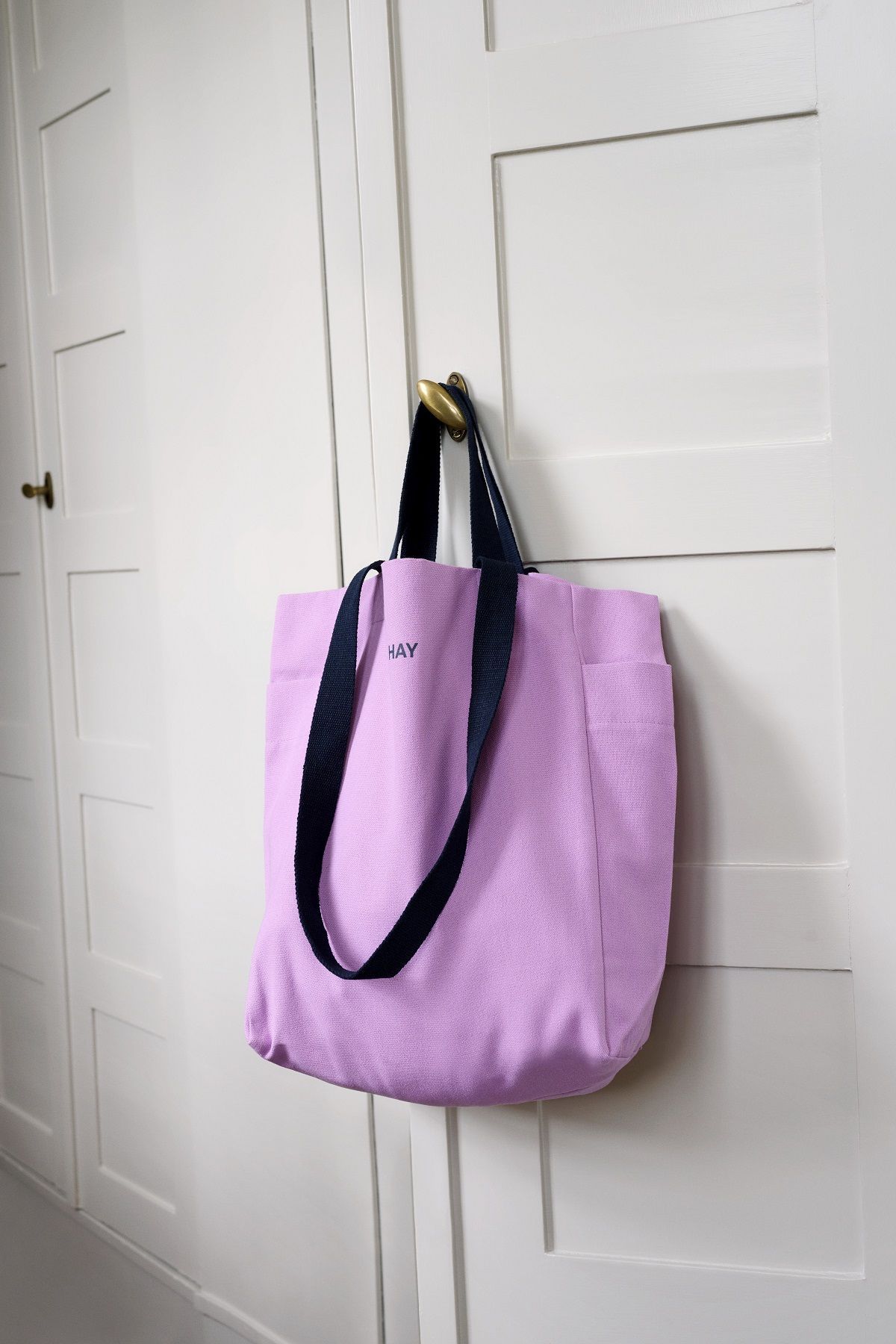 Nahaufnahme der lila Hay Everyday Tote Bag mit dunkelblauen Trägern, hängend an einer weißen Tür.