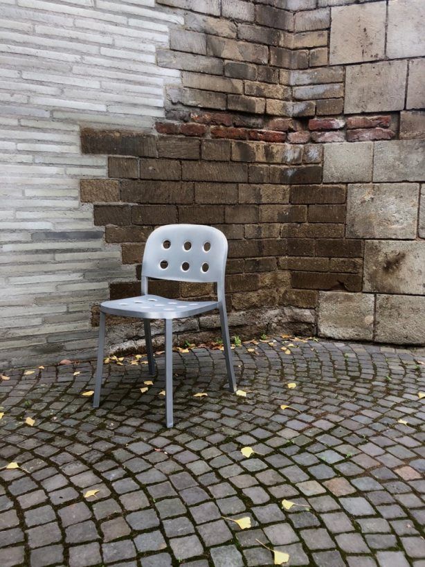 Silberner Emeco 1 Inch Stacking Chair Aluminium Stuhl vor einer Backsteinmauer auf Kopfsteinpflaster mit gelben Blättern.