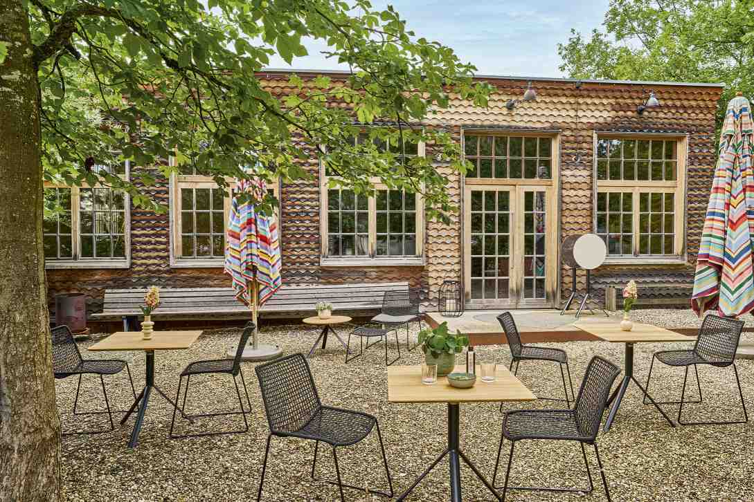 Weishäupl Slope Tischgruppe im Garten: Quadratische Holztische mit schwarzen Stühlen und bunten Sonnenschirmen.