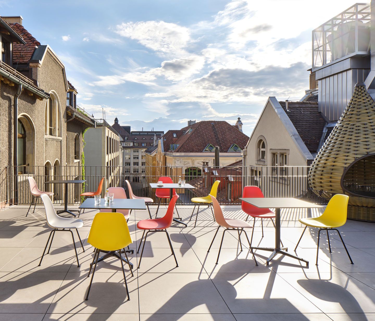 Mehrere farbenfrohe Eames Plastic Side Chairs DSX von Vitra auf einer Dachterrasse mit Tischen.