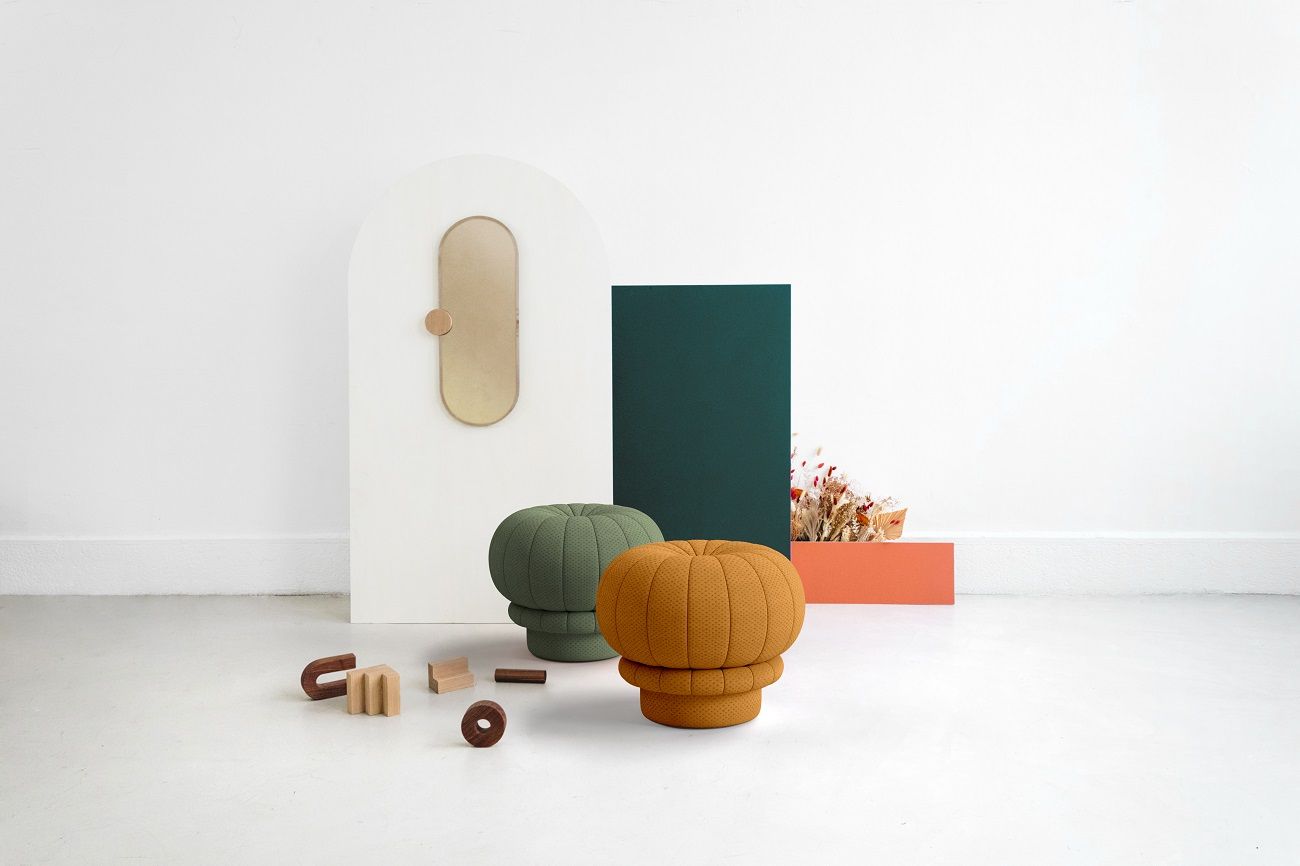 Zwei Claudie Pouf Hocker, einer in Grün, einer in Senfgelb, mit Deko-Elementen im Hintergrund.