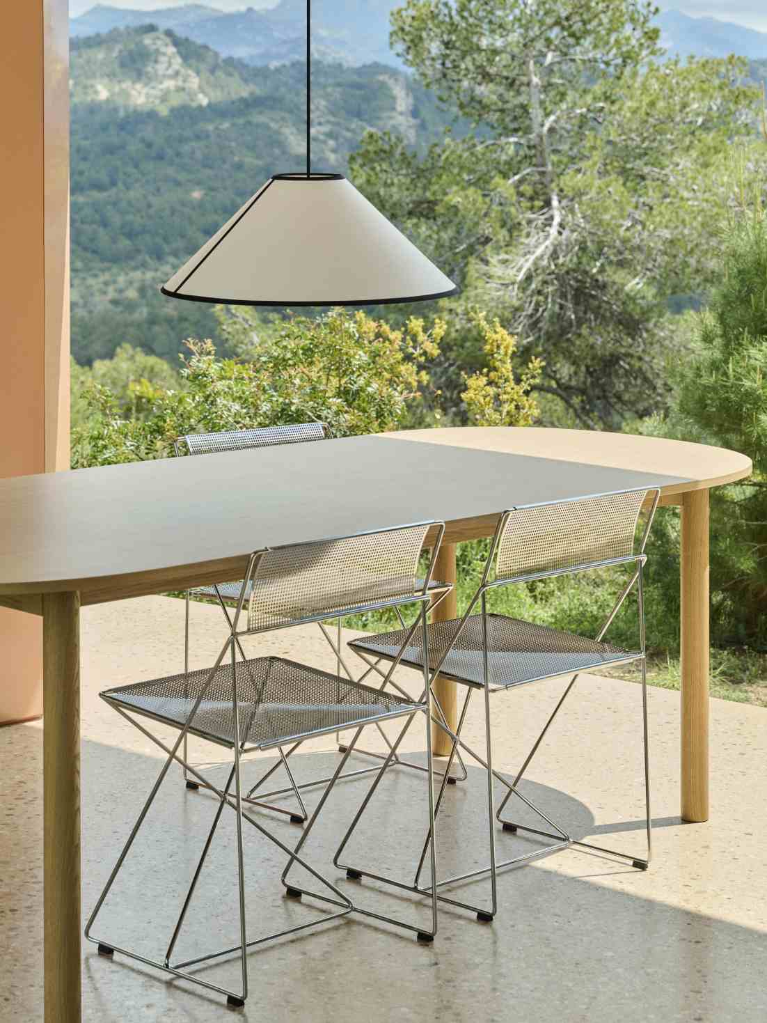 Annex Table Oblong Esstisch Oval Hay mit Metallstühlen und Hängelampe im Freien vor grüner Landschaft.