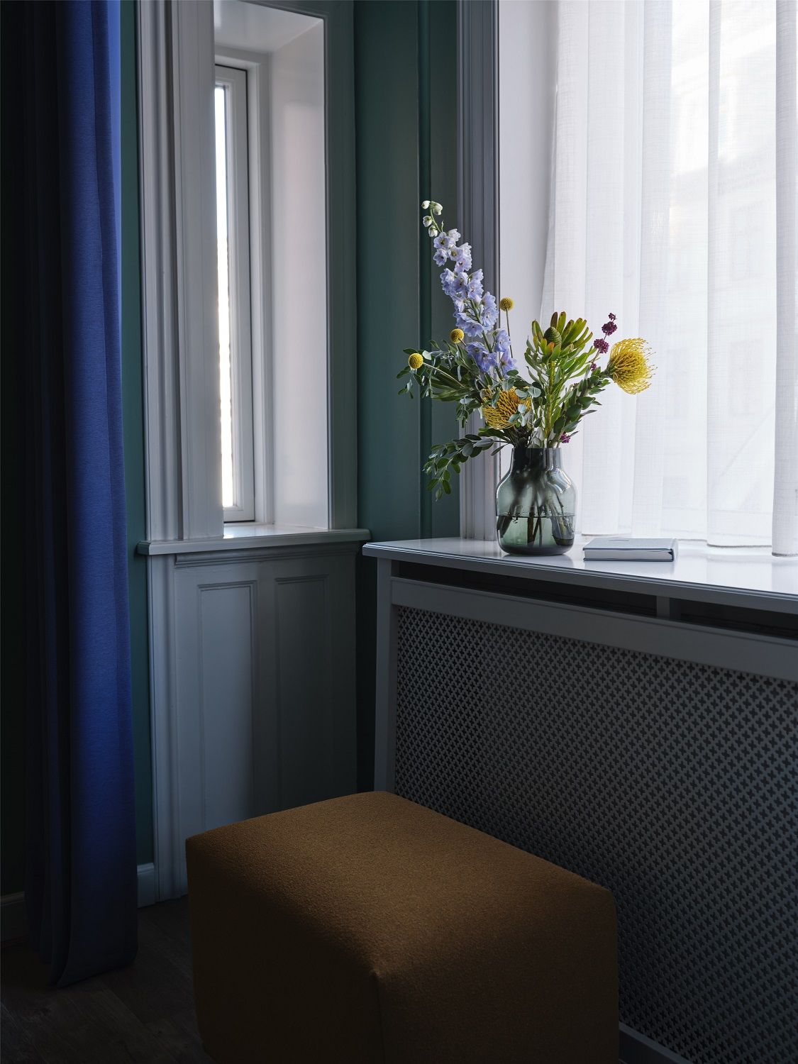 Echo Pouf Hocker von Muuto in Braun vor einem Heizkörper und Blumenstrauß im Interieur.