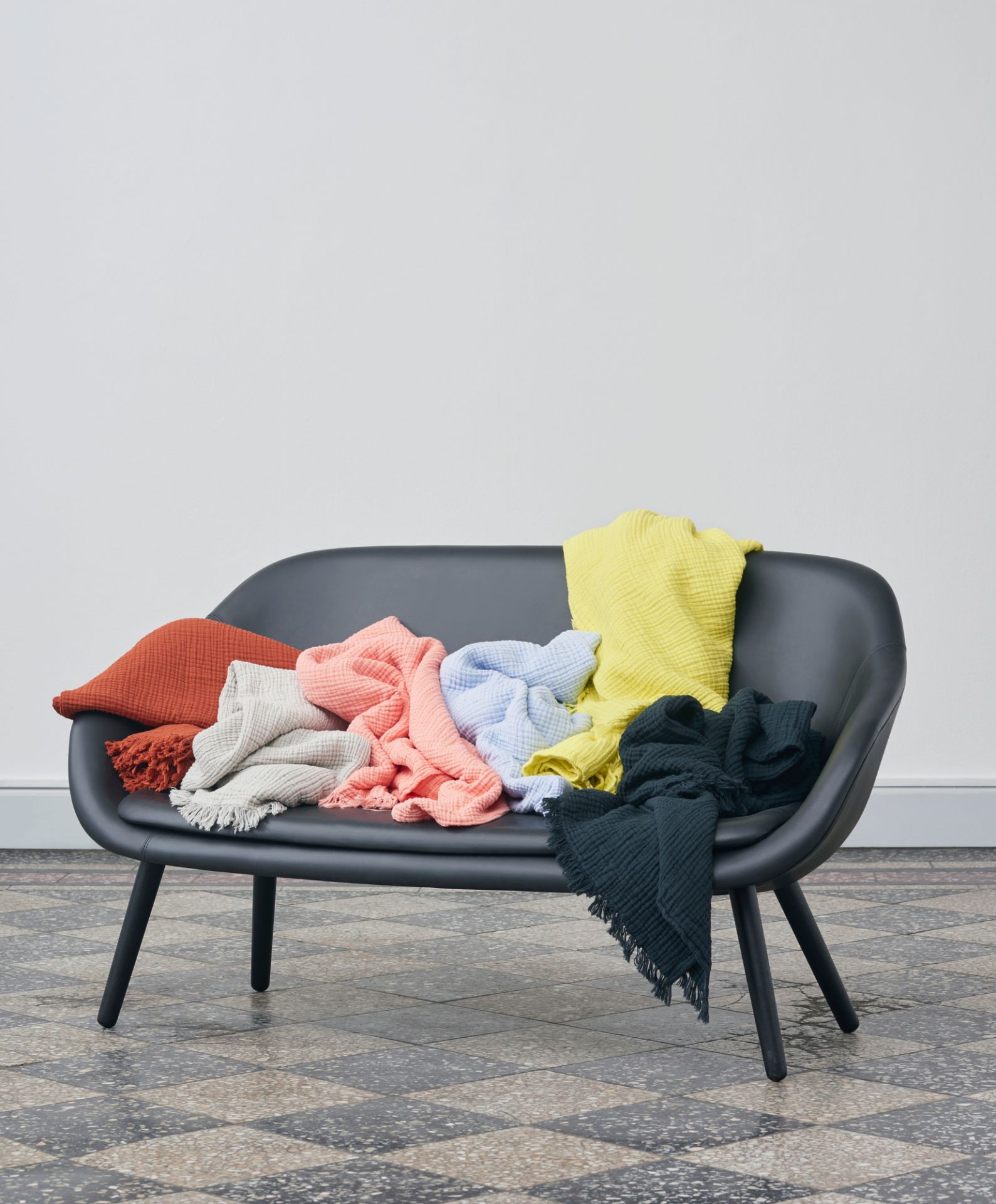 Schwarzes Hay About a Lounge Sofa mit bunten Baumwolldecken auf einem Schachbrettboden vor einer weißen Wand.