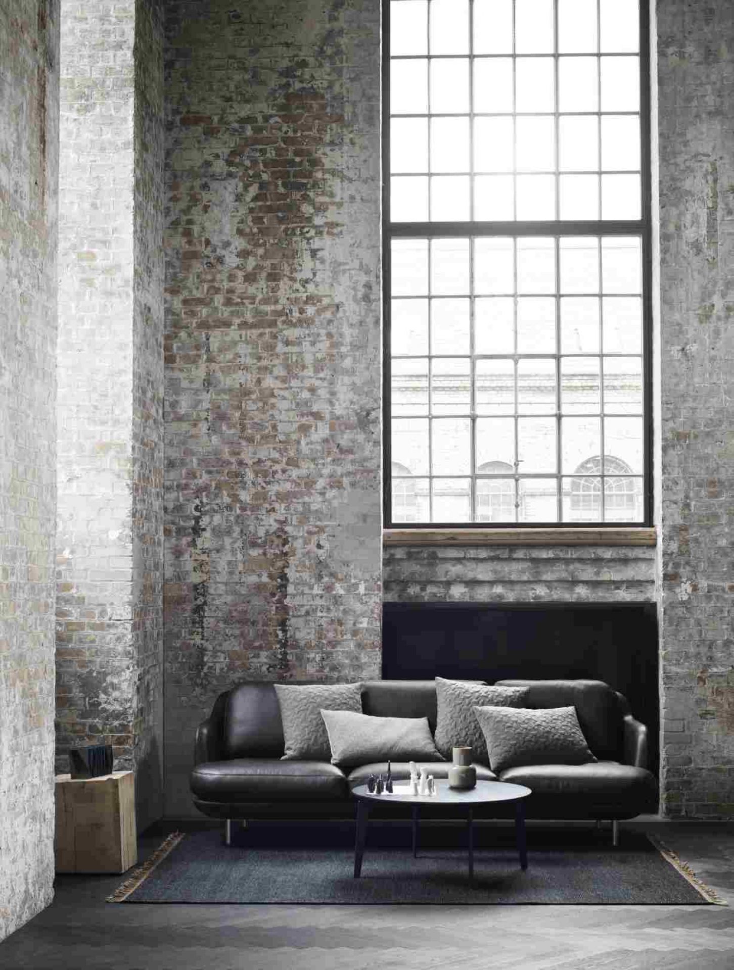 Schwarzes Fritz Hansen Lune Sofa mit grauen Kissen in einem Loft mit Backsteinwand und großem Fenster.