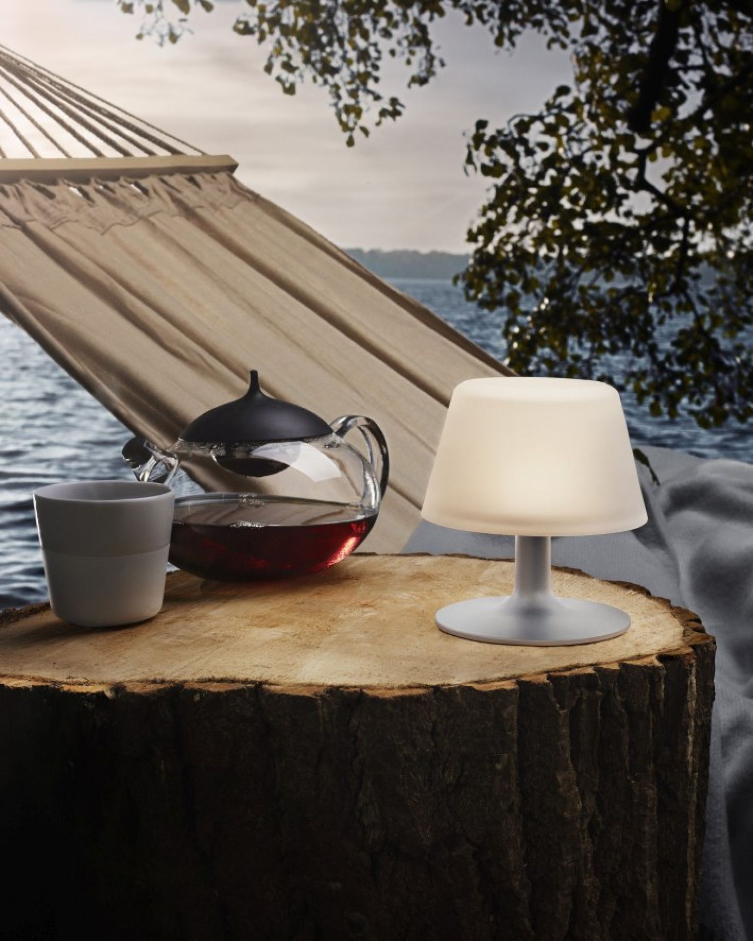 Sunlight Tischleuchte von Eva Solo auf Baumstumpf mit Tee und Hängematte im Hintergrund.