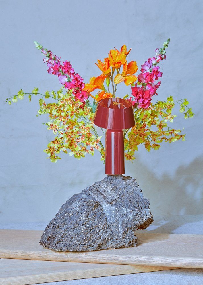 Marset Bicoca Tischleuchte in Rot auf Stein, umgeben von bunten Blumen. Akkuleuchte für Innen.