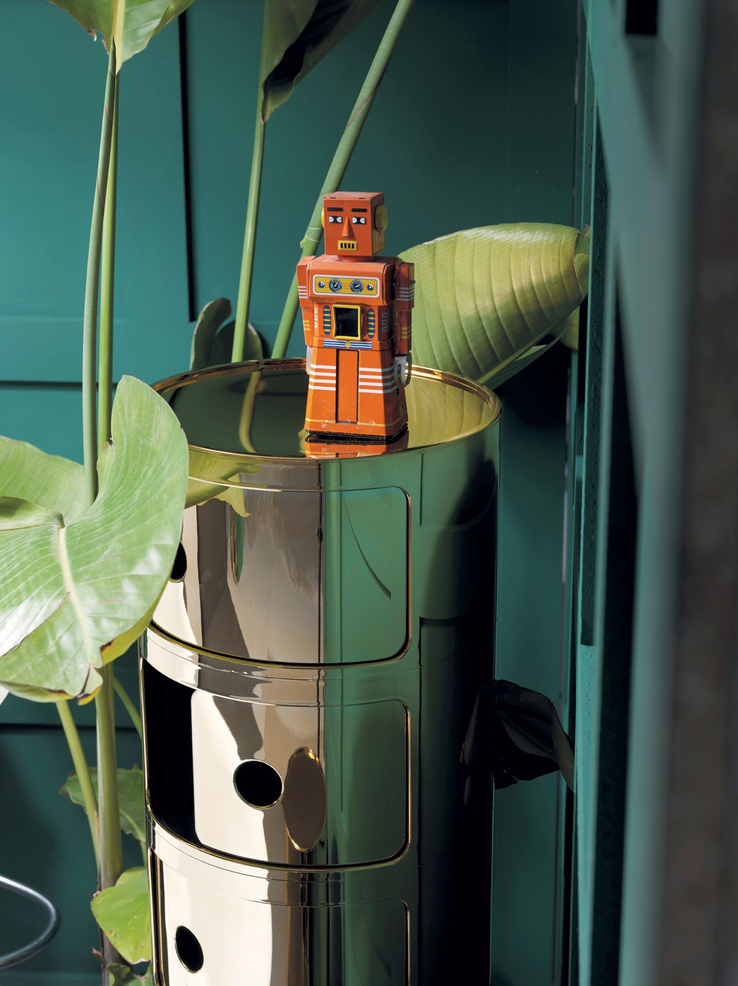 Goldener Componibili 3er Container von Kartell mit Spielzeugroboter und grünen Pflanzen vor grüner Wand.