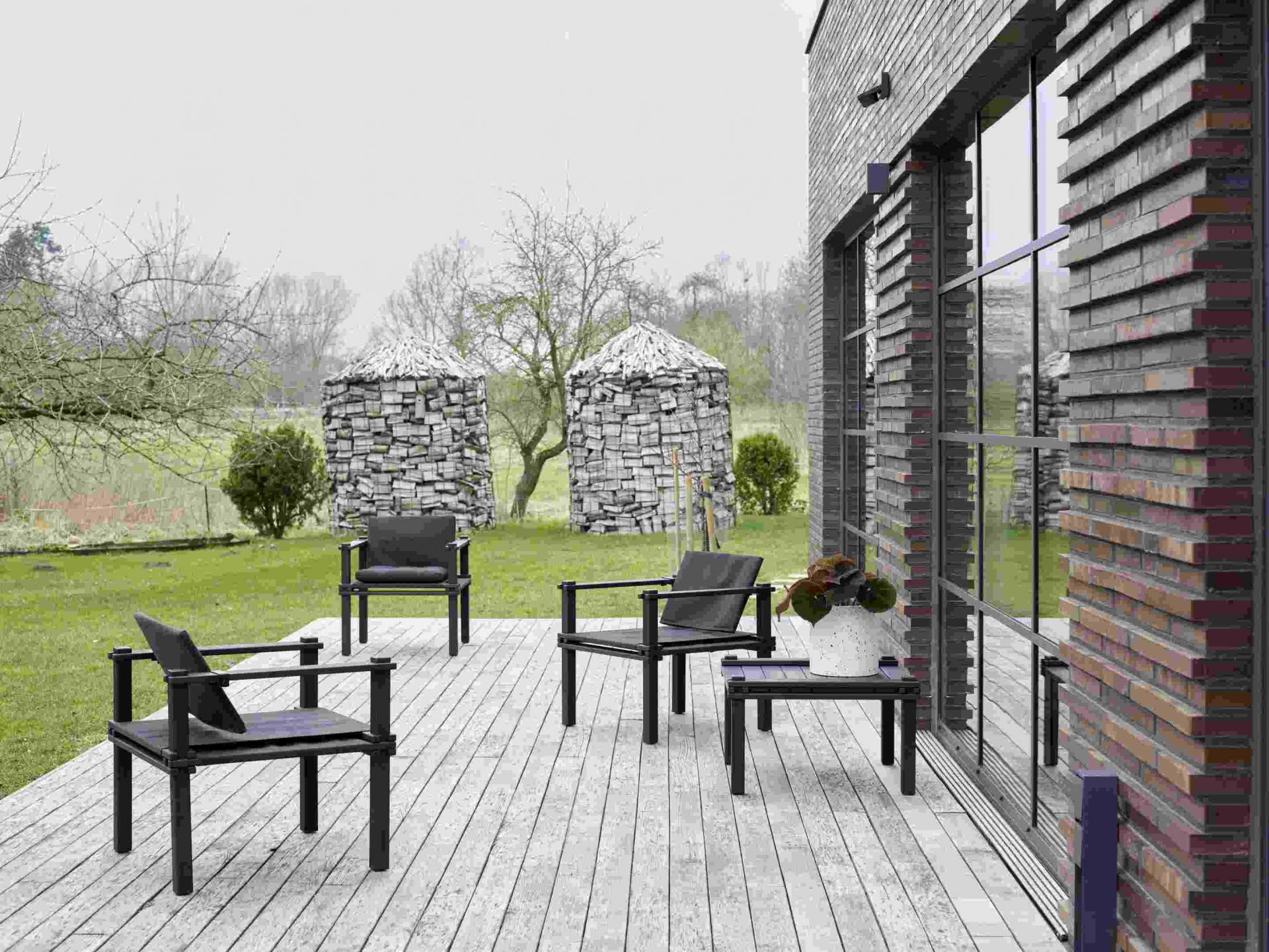 Drei Farmer Sessel Outdoor Cor auf einer Holzterrasse mit Blick auf einen Garten und ein modernes Haus.