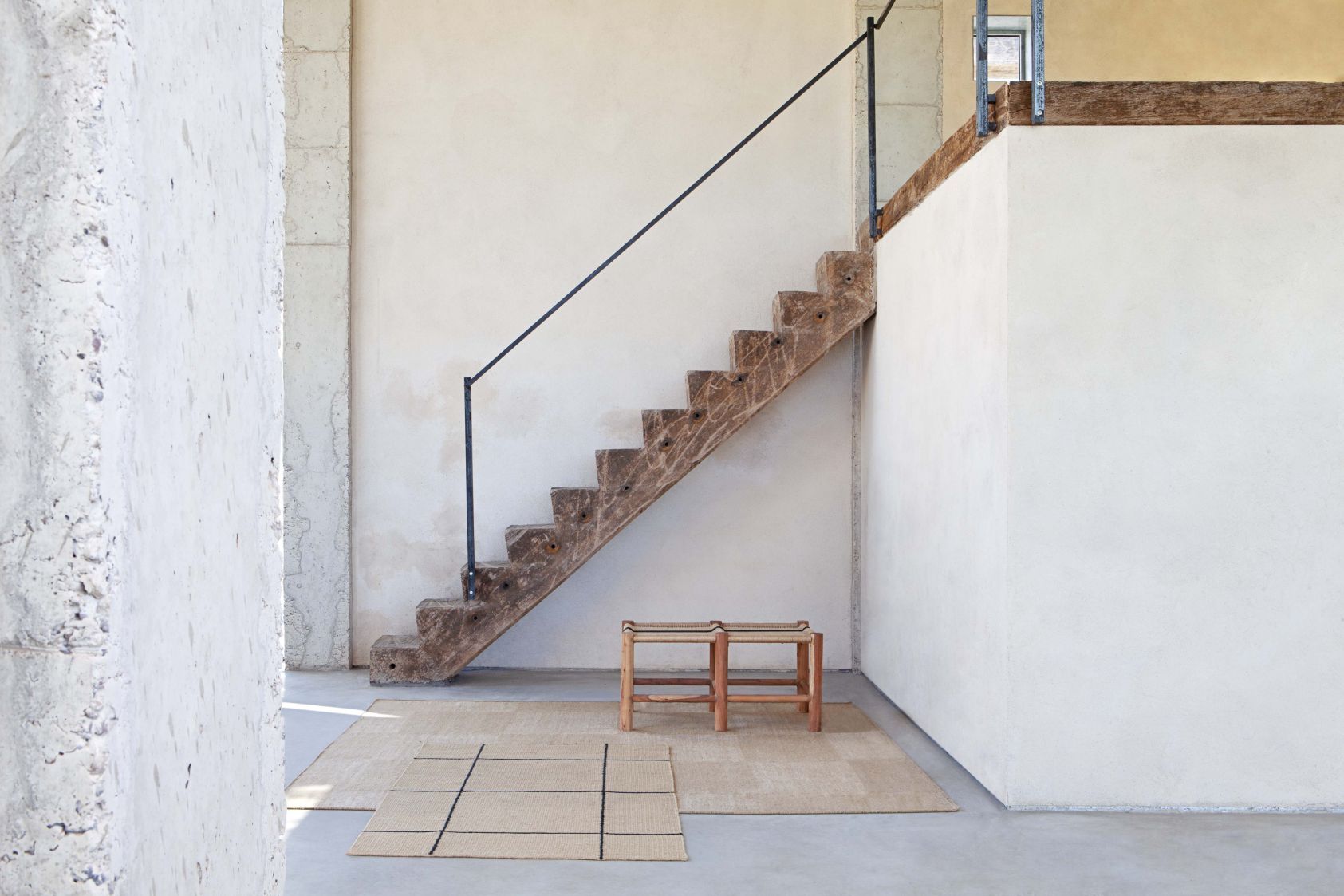 Roots Double Stool Bank Gan aus Holz und Rattan vor einer Treppe und einem Teppich.