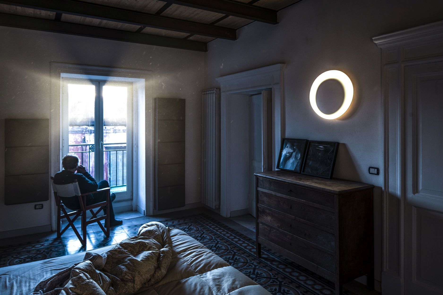 Bahia Mini Wandleuchte von Foscarini in einem Schlafzimmer mit Bett und Person am Fenster.