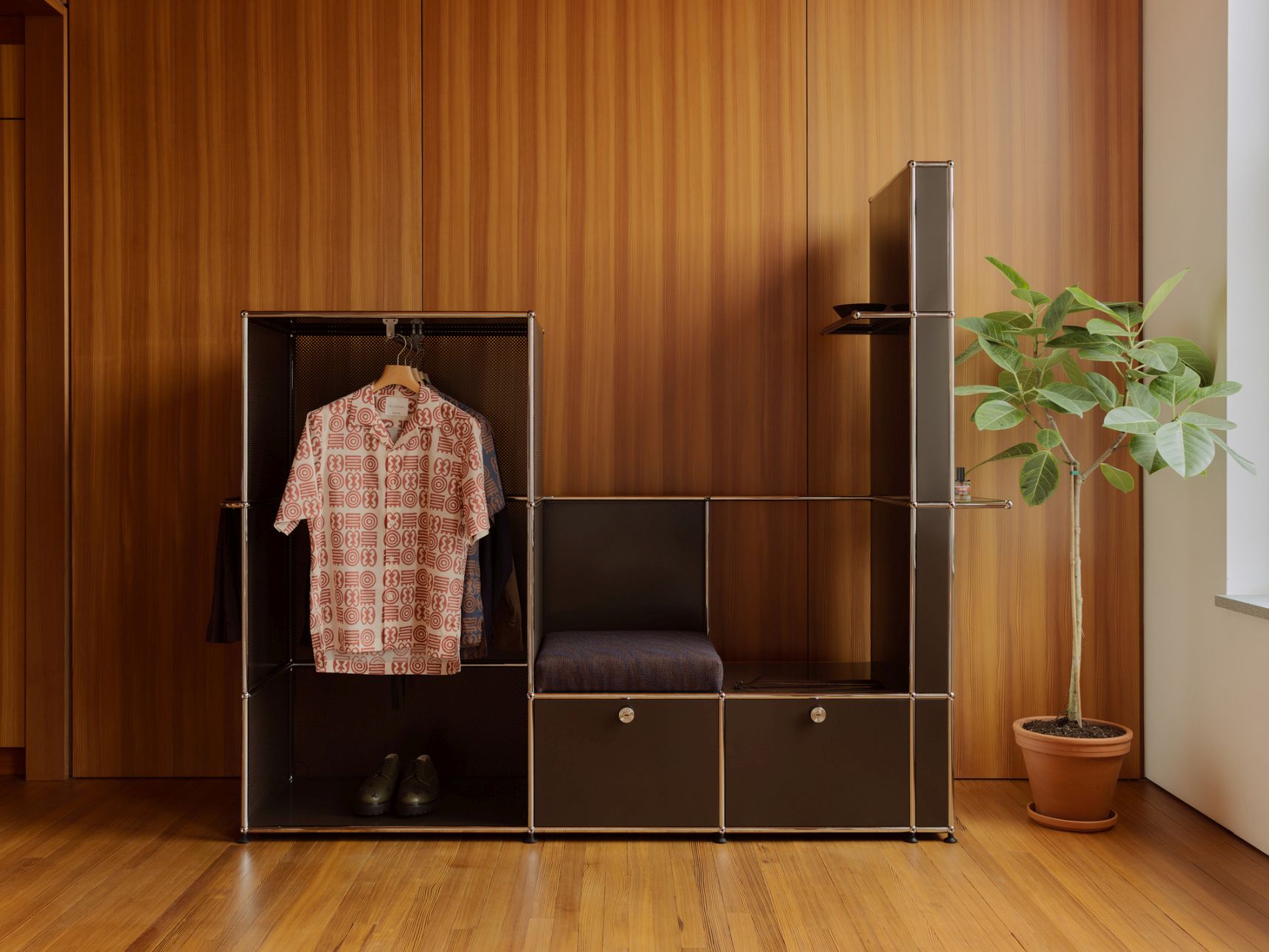 USM Haller Okinka Garderobe mit Kleiderstange, Sitzbank, Ablagefächern und Zimmerpflanze vor Holzwand.
