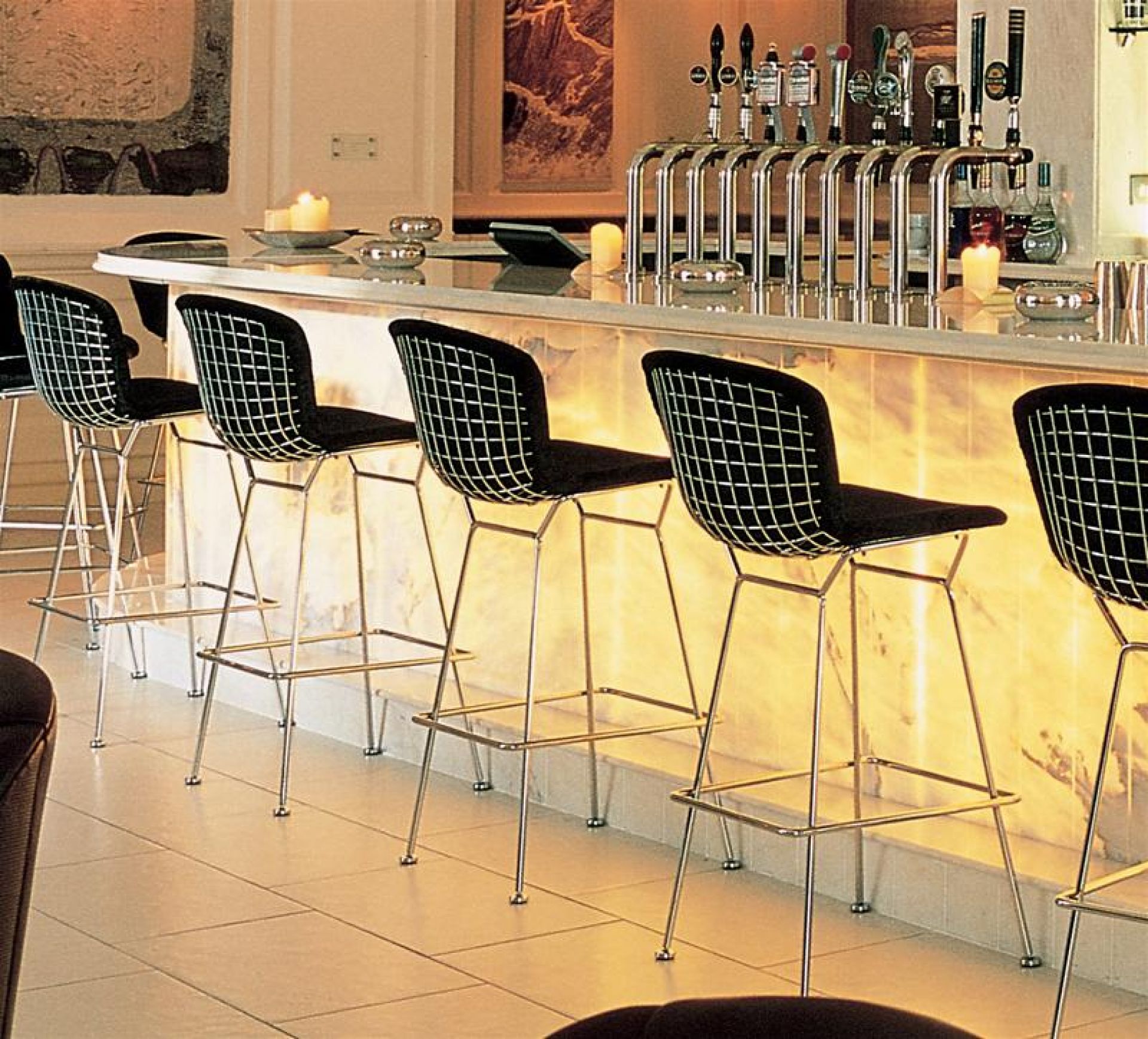 Bertoia Barhocker von Knoll International, Design-Barhocker mit schwarzer Sitzfläche und verchromtem Stahlgestell vor beleuchteter Bar.