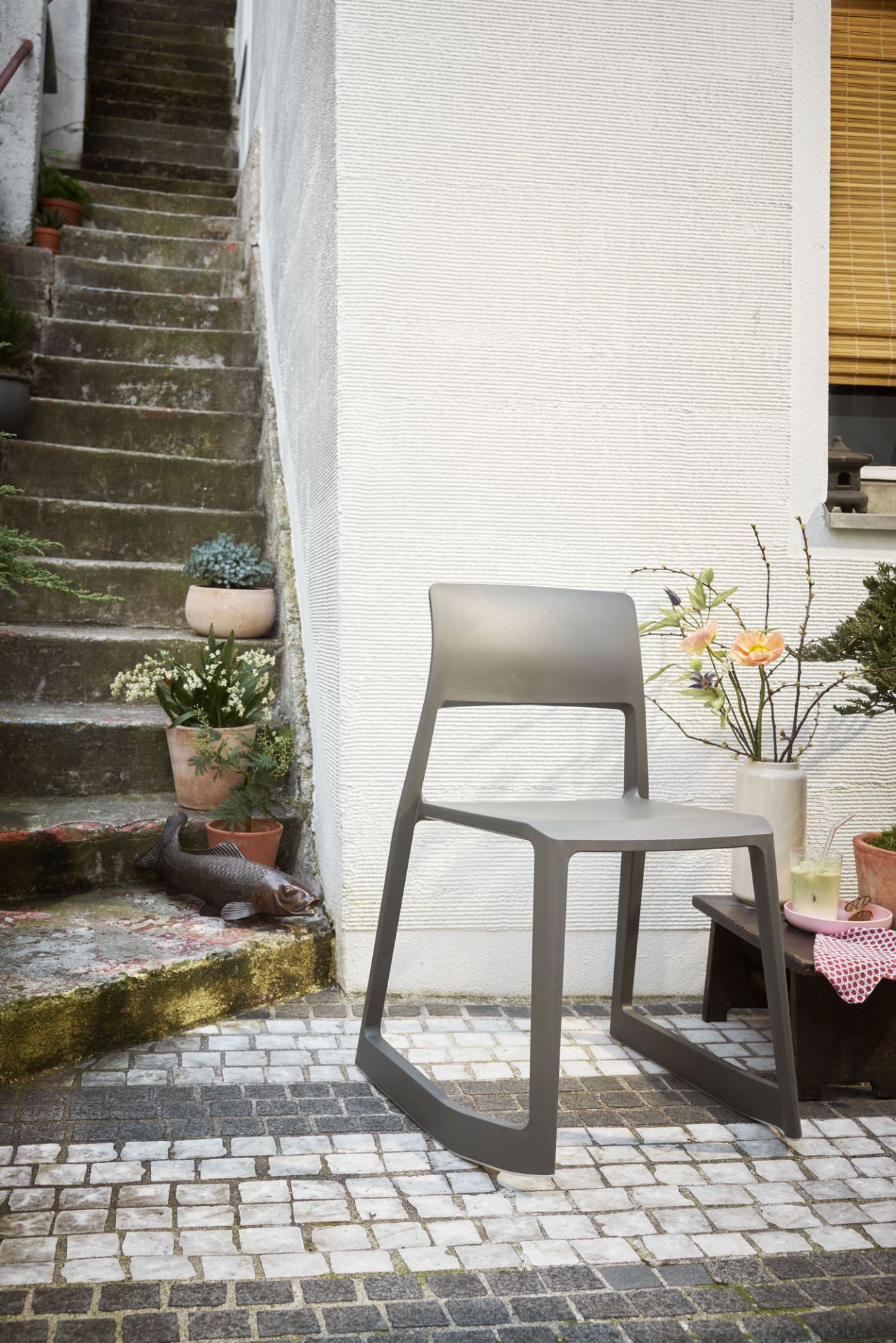 Vitra Tip Ton RE Stuhl in Soft Grey vor weißer Wand und Treppe mit Pflanzen.