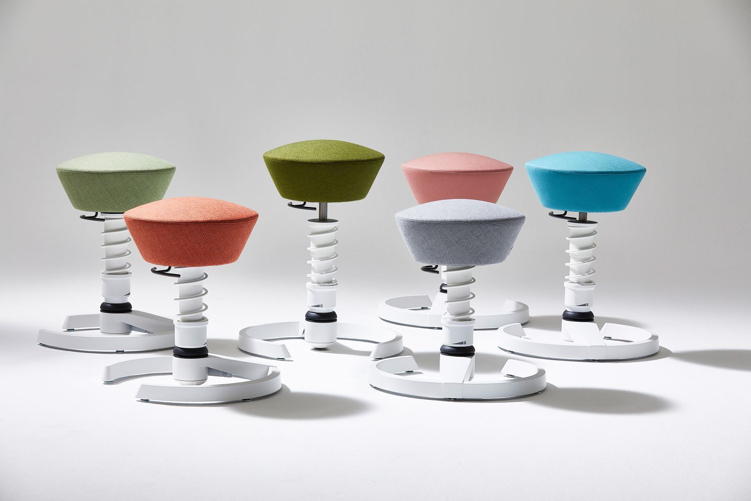 Mehrere farbige, moderne Stehhocker mit weißem Gestell und rundem Fuß für ergonomisches Sitzen.