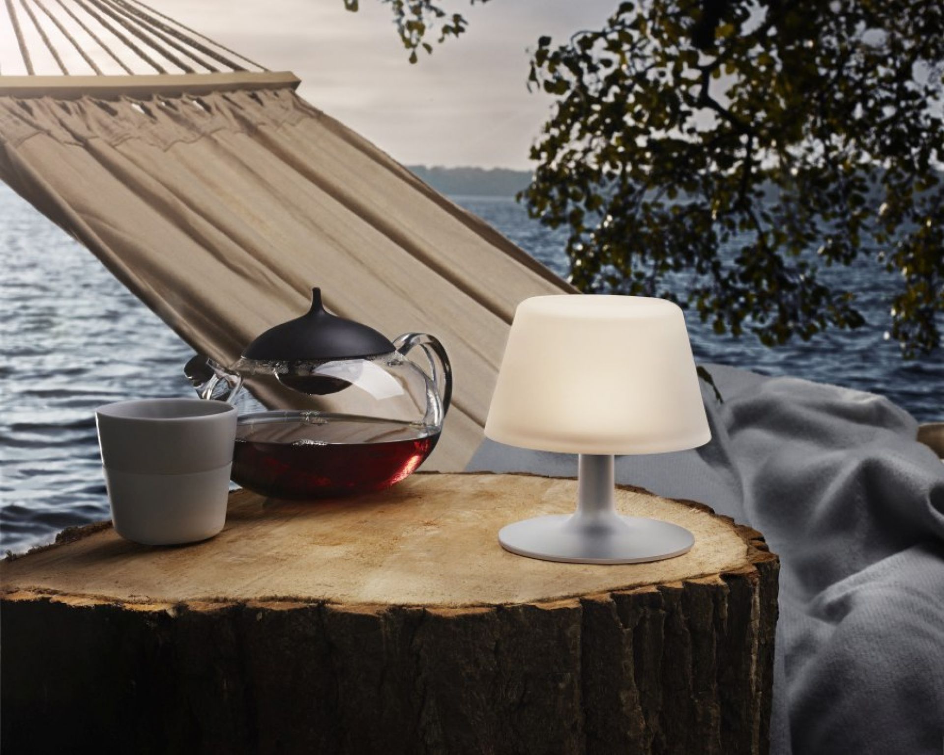 Sunlight Tischleuchte von Eva Solo auf Baumstumpf mit Tee und Hängematte im Hintergrund.
