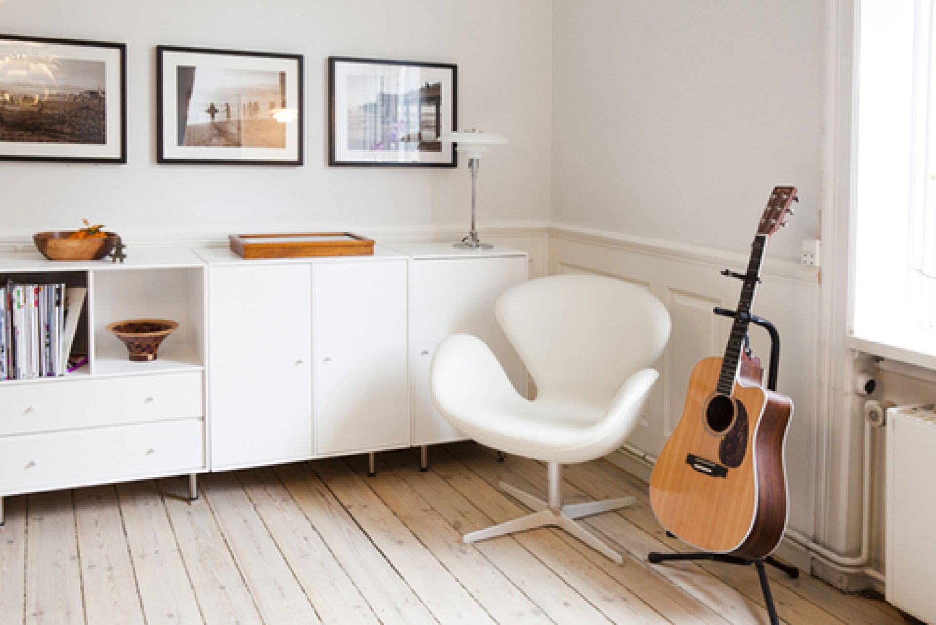 Heller Raum mit weißem Fritz Hansen Schwan Sessel, Gitarre und weißen Möbeln unter gerahmten Bildern.
