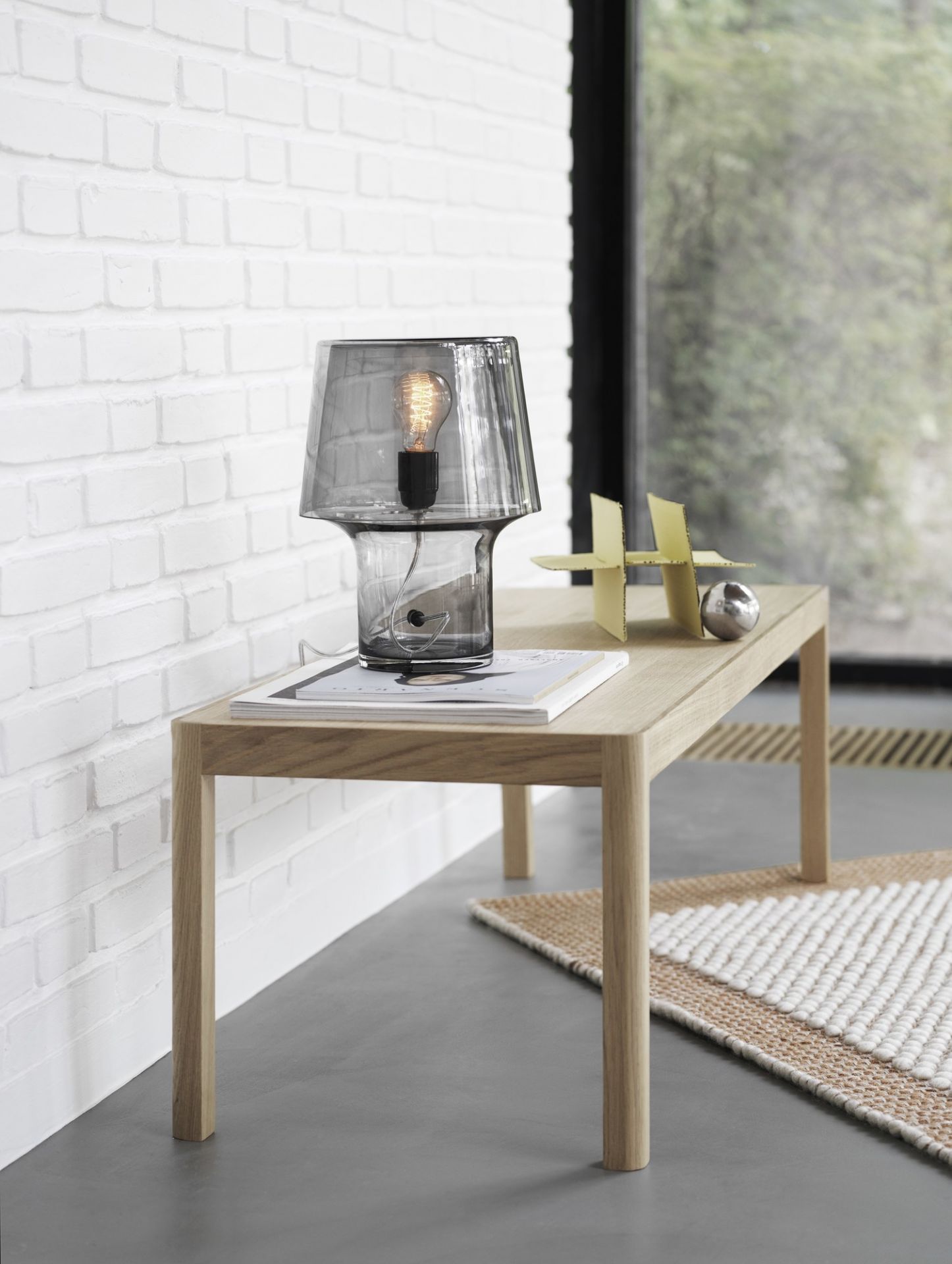 Nahaufnahme des Muuto Pebble Rug Teppichs, teilweise unter einem Holztisch sichtbar, mit grauer Lampe.